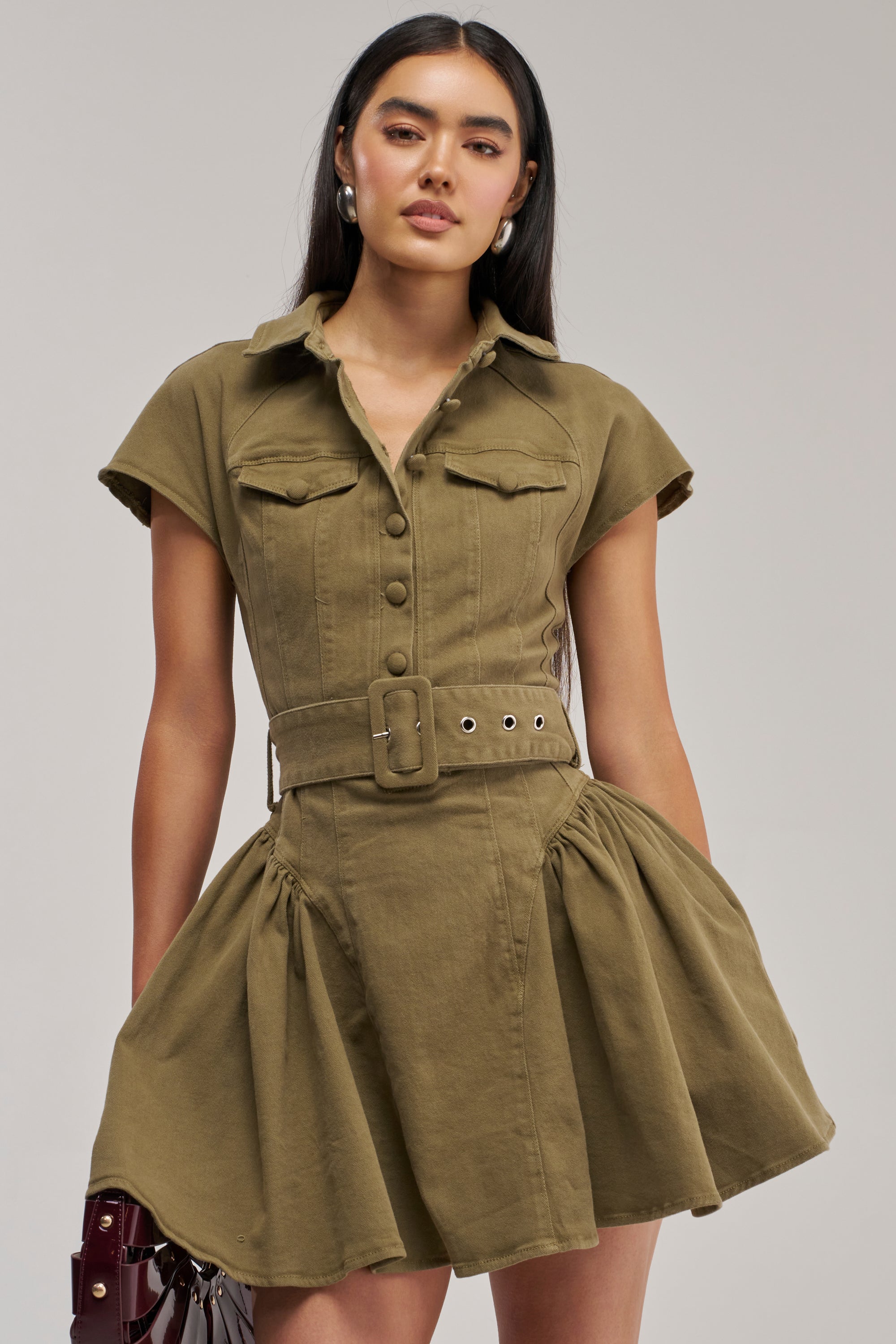 A woman with long dark hair stands against a plain light background wearing the SADIE BUTTON UP MINI DRESS IN OLIVE DENIM, a short-sleeved mini dress featuring a belted waist, chest pockets, an olive-green hue, button-down front, and flared skirt.