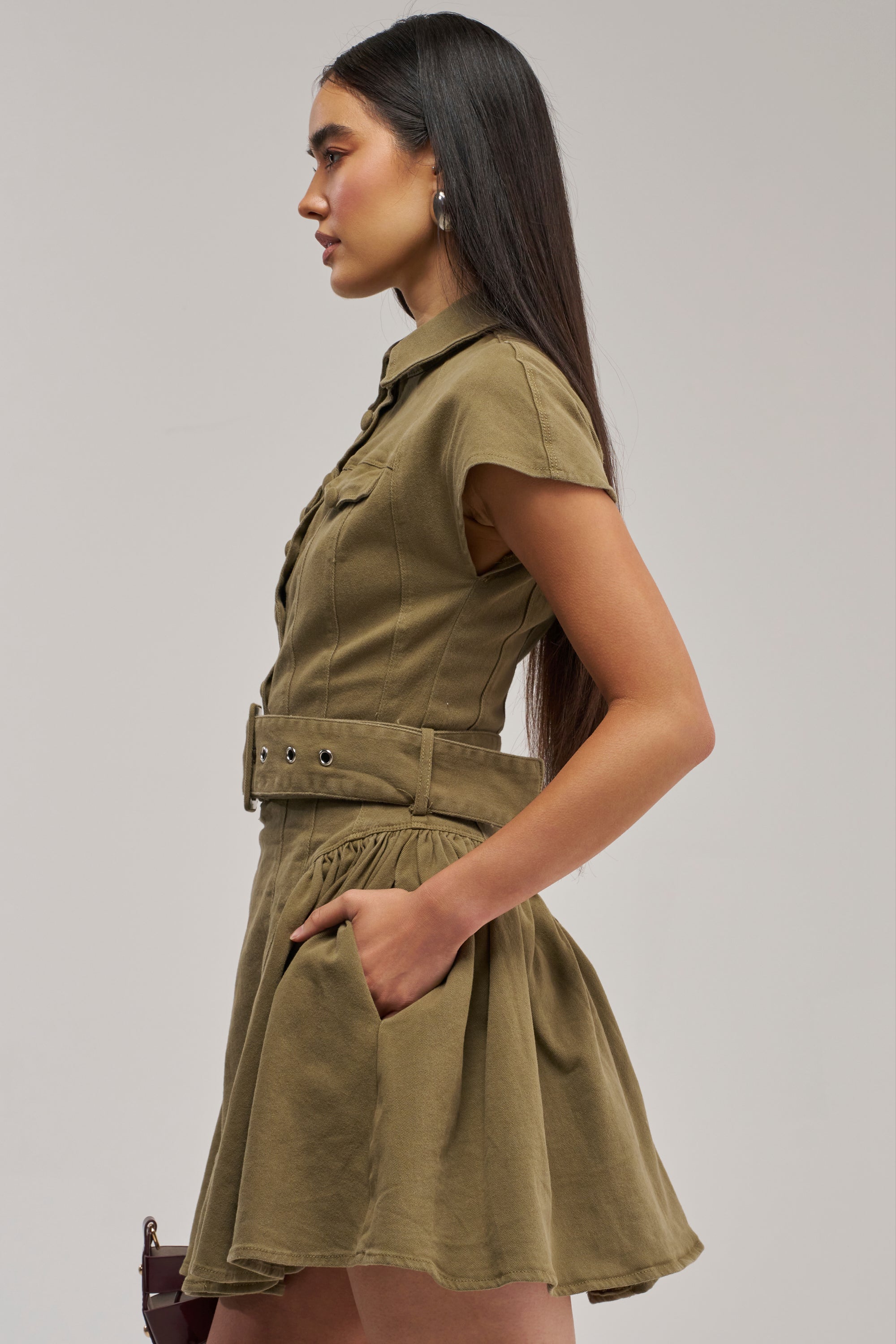 A woman in profile wearing the SADIE Button Up Mini Dress in Olive Denim, a short-sleeve belted dress with a flared skirt, stands with one hand in her pocket against a plain background.