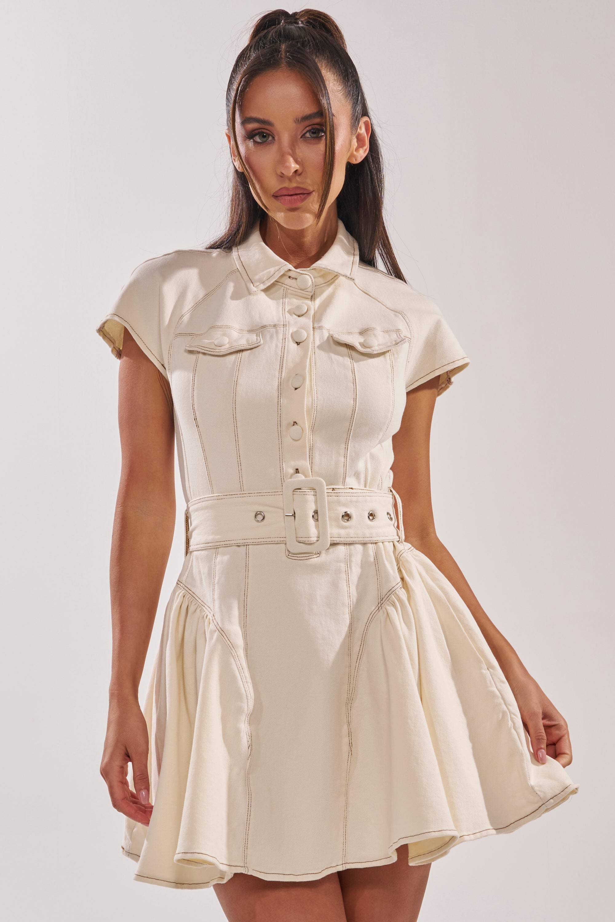 A woman models the SADIE BUTTON UP DENIM MINI DRESS IN WHITE—a short-sleeved, belted mini dress with a flared skirt, button-down front, and two chest pockets—standing against a plain light background. 1