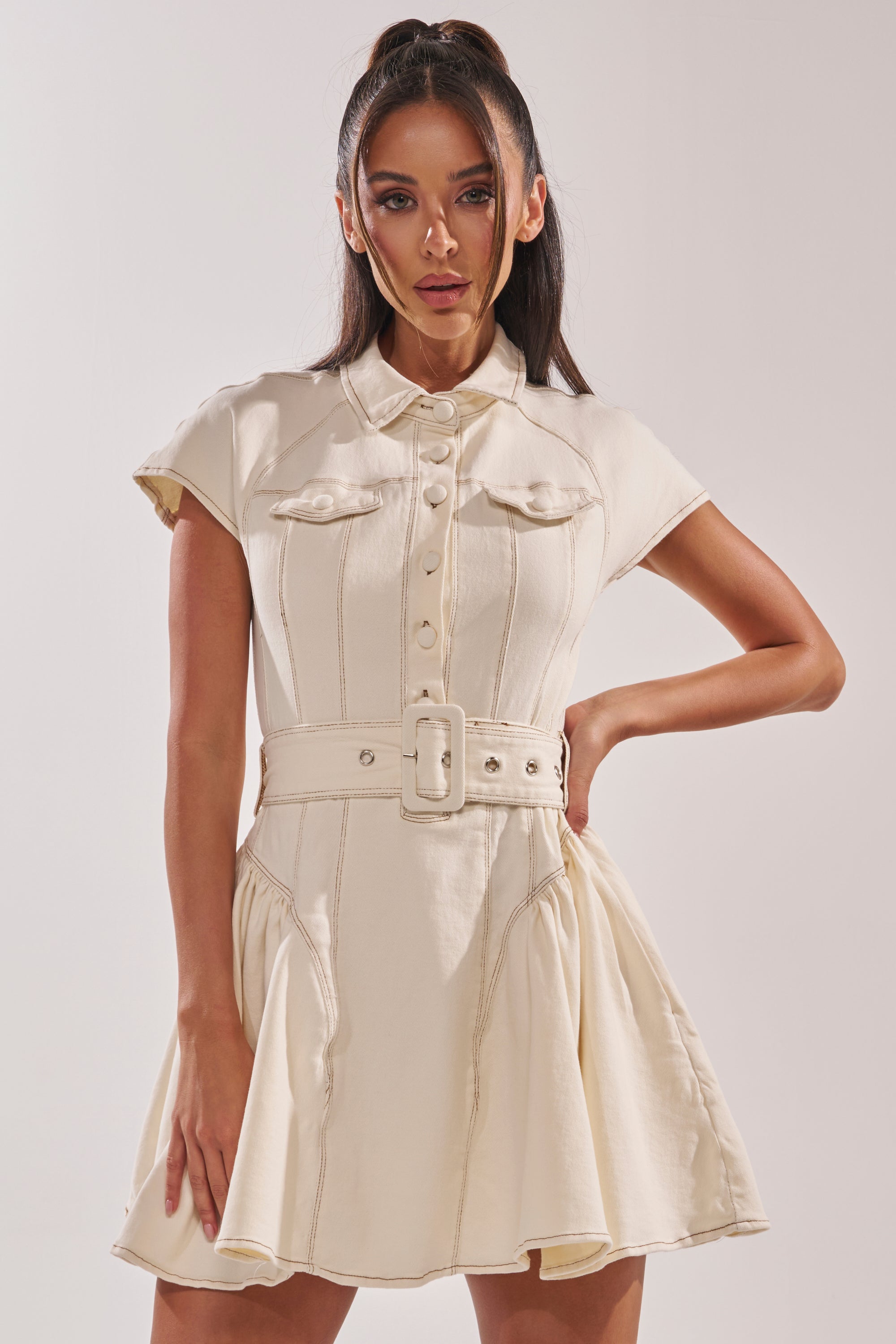 A woman models the SADIE BUTTON UP DENIM MINI DRESS IN WHITE, featuring short sleeves, a button-down front, chest pockets, and a flared skirt. She wears her long dark hair in a high ponytail against a plain background. 2