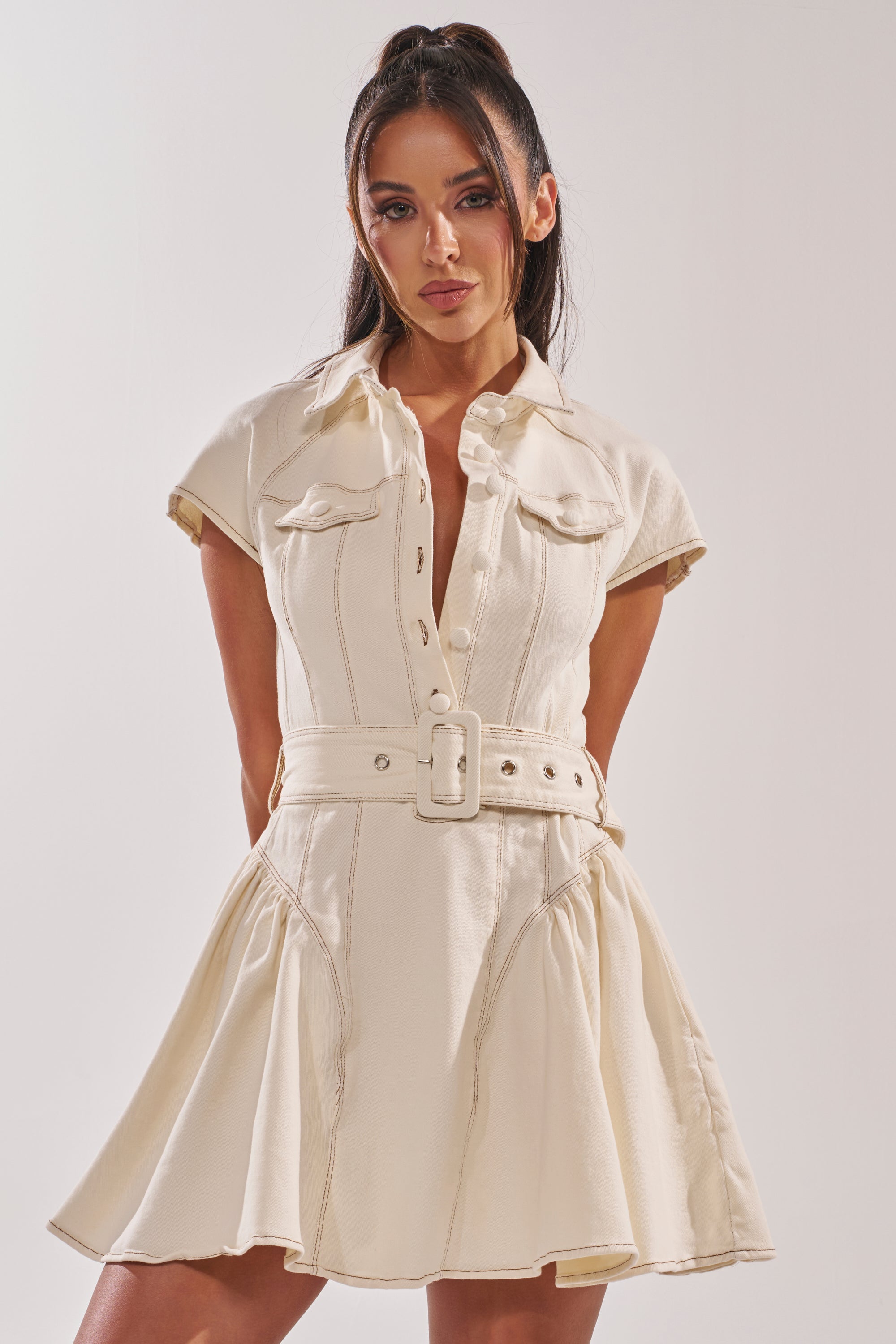A woman stands against a plain light background, looking directly at the camera, wearing the SADIE BUTTON UP DENIM MINI DRESS IN WHITE with short sleeves, a belted waist, and a flared mini skirt. 3