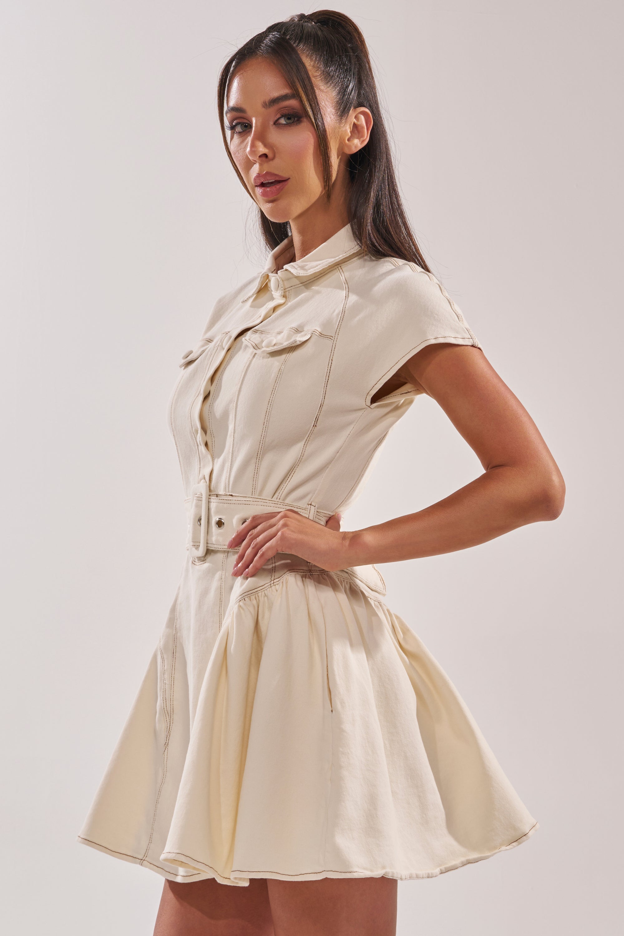 A woman with long dark hair in a high ponytail models the SADIE BUTTON UP DENIM MINI DRESS IN WHITE, featuring short sleeves, a collared neckline, belted waist, and flared skirt, posing sideways against a plain light background. 4