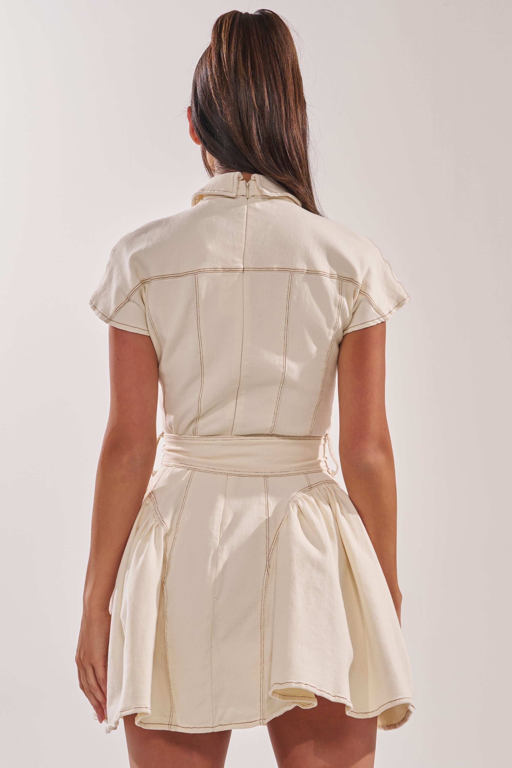 A woman with long brown hair in a high ponytail is shown from the back, wearing the SADIE BUTTON UP DENIM MINI DRESS IN WHITE, which features visible seam details and a flared skirt against a plain background. 5