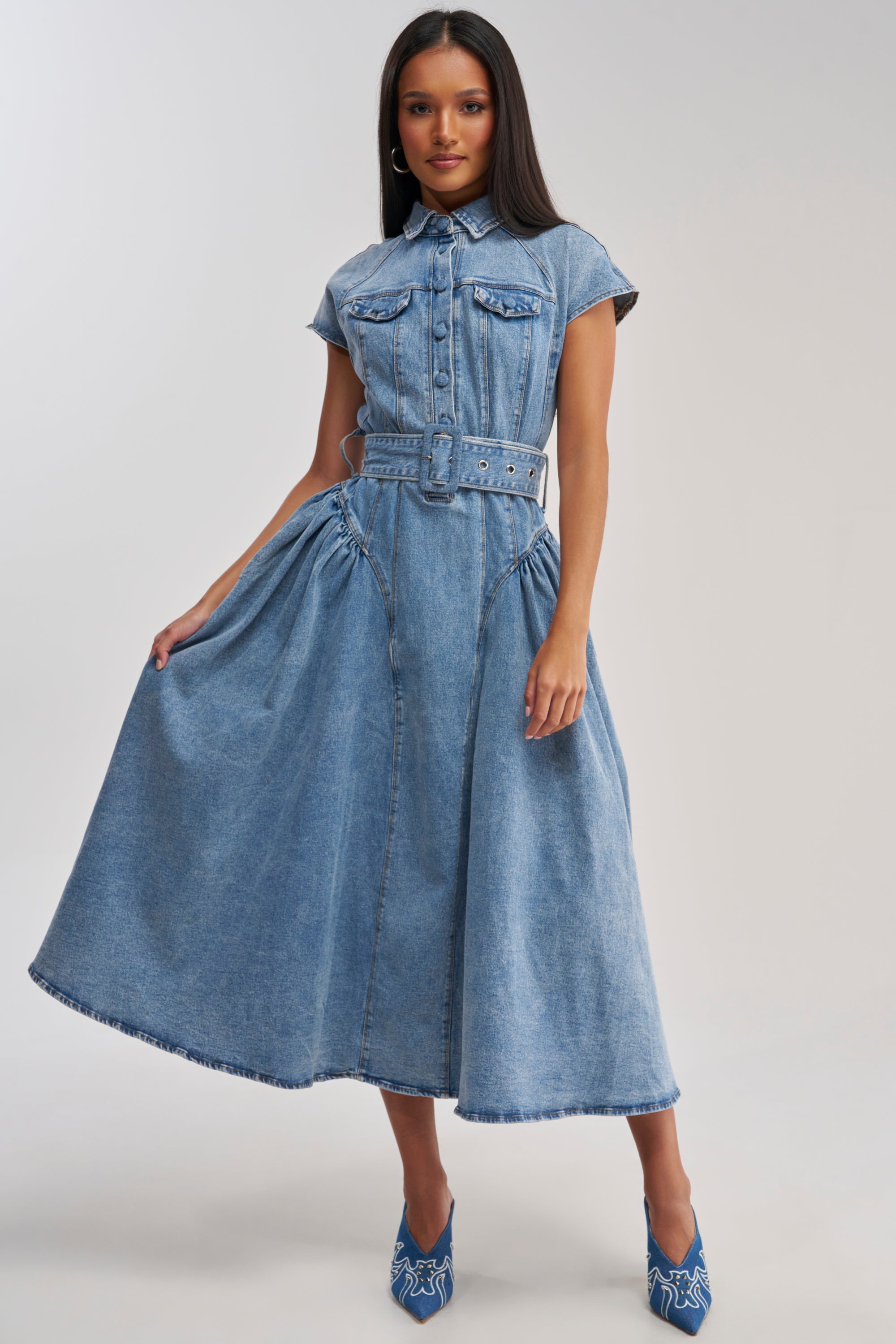 A woman poses in the MEET THE PARENTS BELTED DENIM MIDI DRESS, featuring a collar, short sleeves, front pockets, pleats, and a belt. She pairs it with blue heels with white patterns against a plain light background.