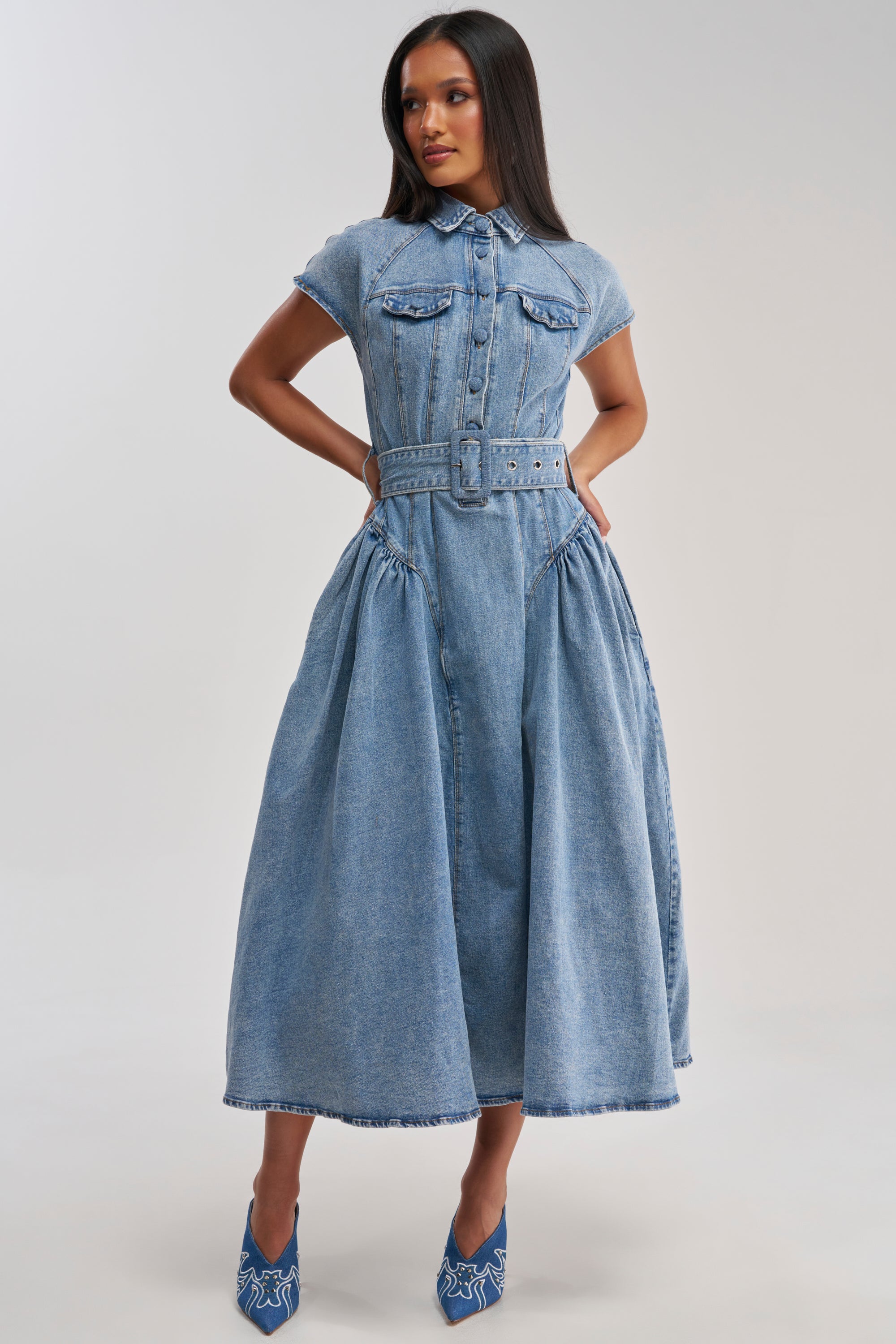 A woman models the MEET THE PARENTS BELTED DENIM MIDI DRESS, featuring a collared neckline, short sleeves, chest pockets, belted waist, and a flared skirt. She pairs it with blue patterned heels against a light background.