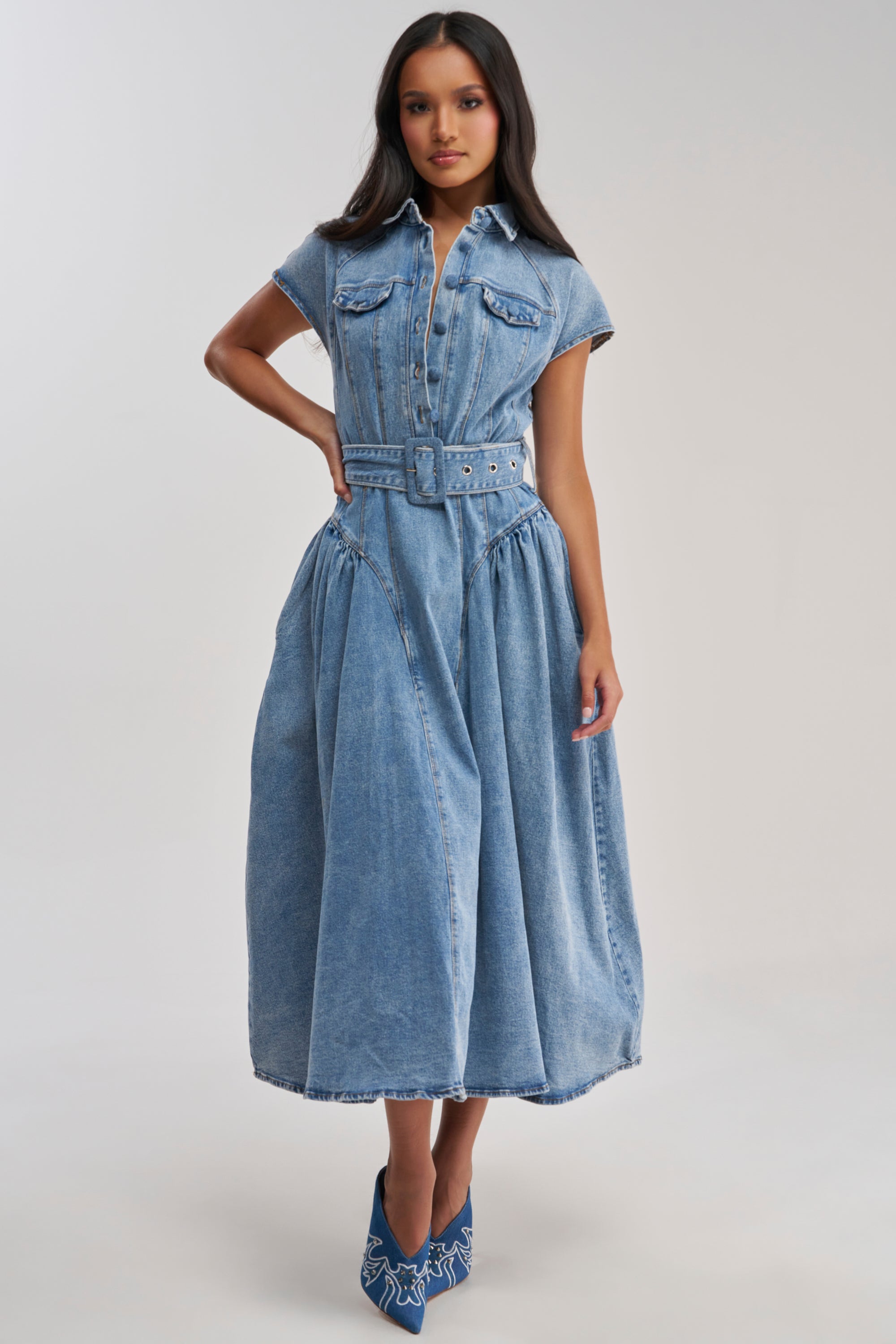 A woman wears the MEET THE PARENTS BELTED DENIM MIDI DRESS—collared, short-sleeved, button-up front, belted waist, flared skirt, and chest pockets—paired with blue pointed-toe shoes featuring a white pattern.