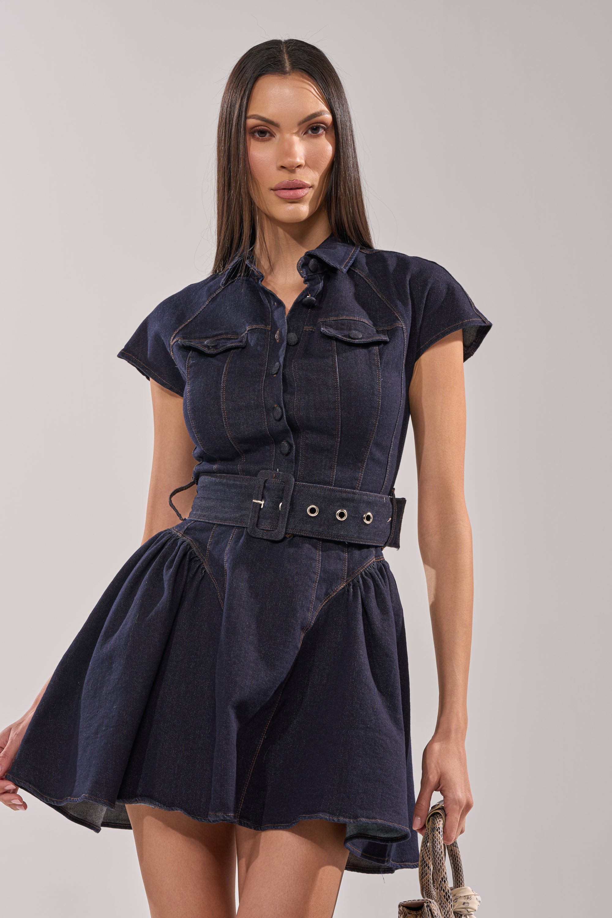 A woman with long straight hair wears the SADIE BUTTON UP DENIM MINI DRESS—a short-sleeved mini dress with a belted waist, button-up front, and skater silhouette. She holds a woven bag and stands against a plain light background. 1