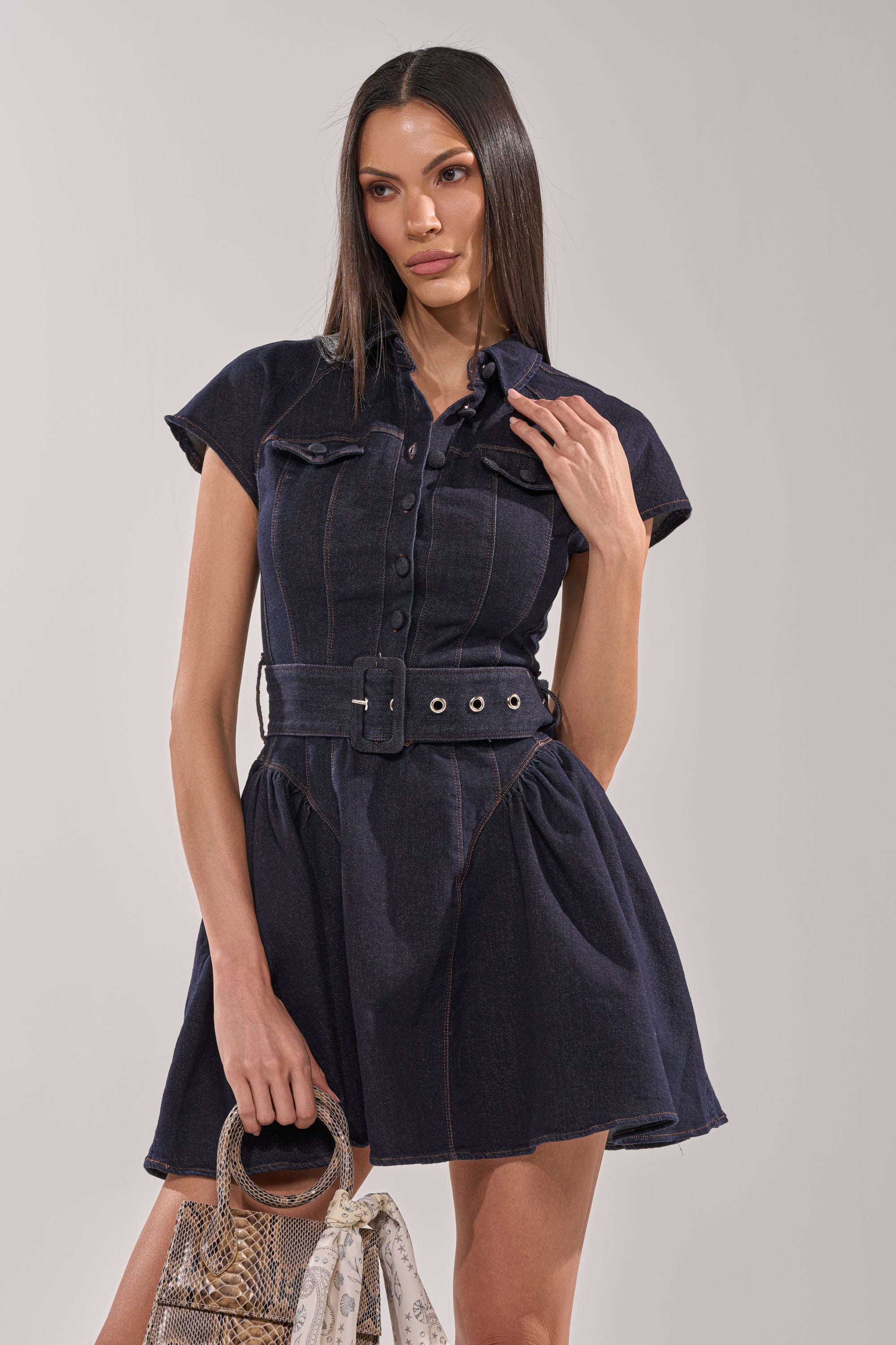 A woman with long straight hair wears the SADIE BUTTON UP DENIM MINI DRESS, which features a belted waist and short sleeves. She holds a handbag with a scarf accent and poses against a plain, light background. 2