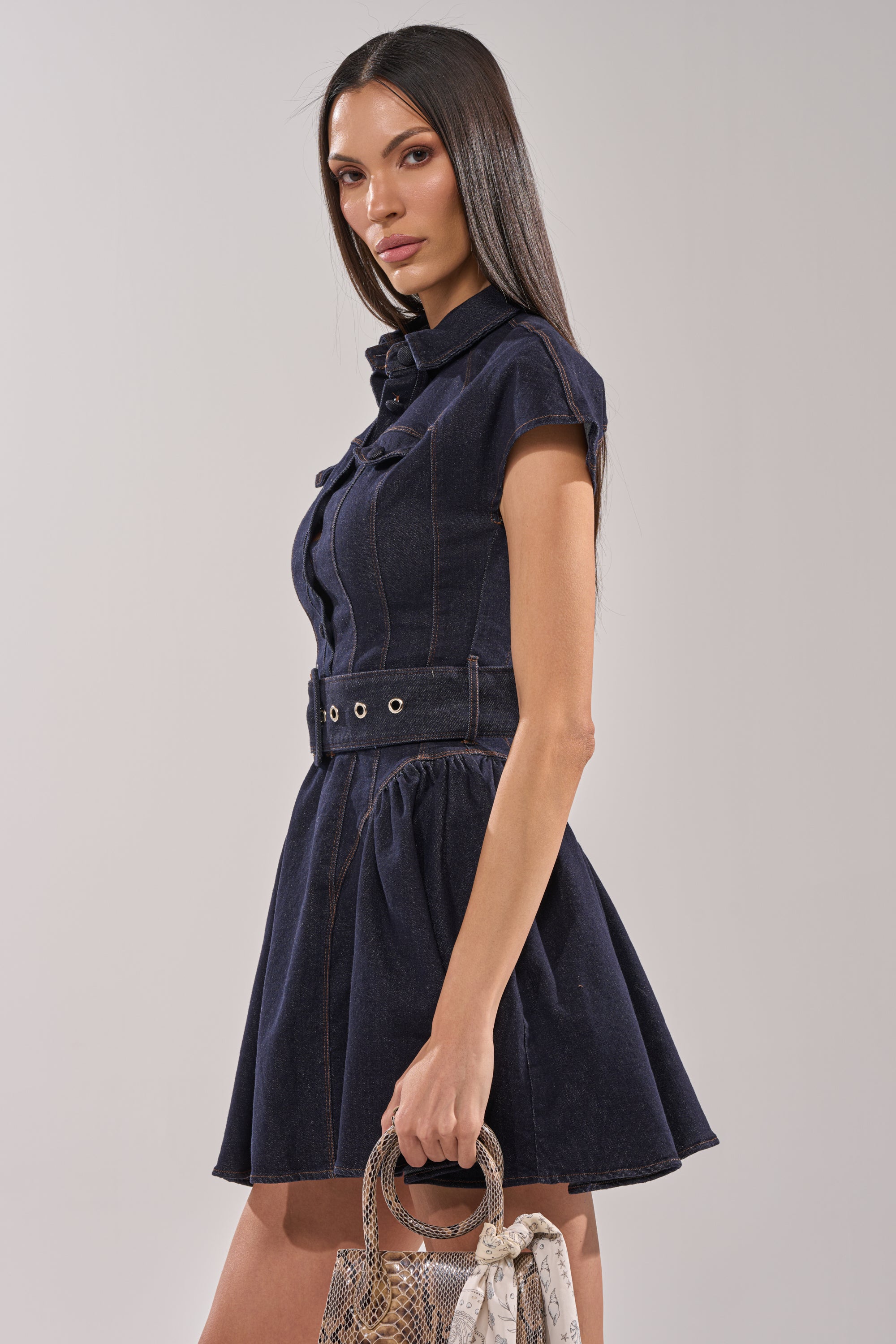 A woman wears the SADIE BUTTON UP DENIM MINI DRESS with a belted waist and skater silhouette, holding a small snakeskin-patterned handbag while posing against a plain light background. 3