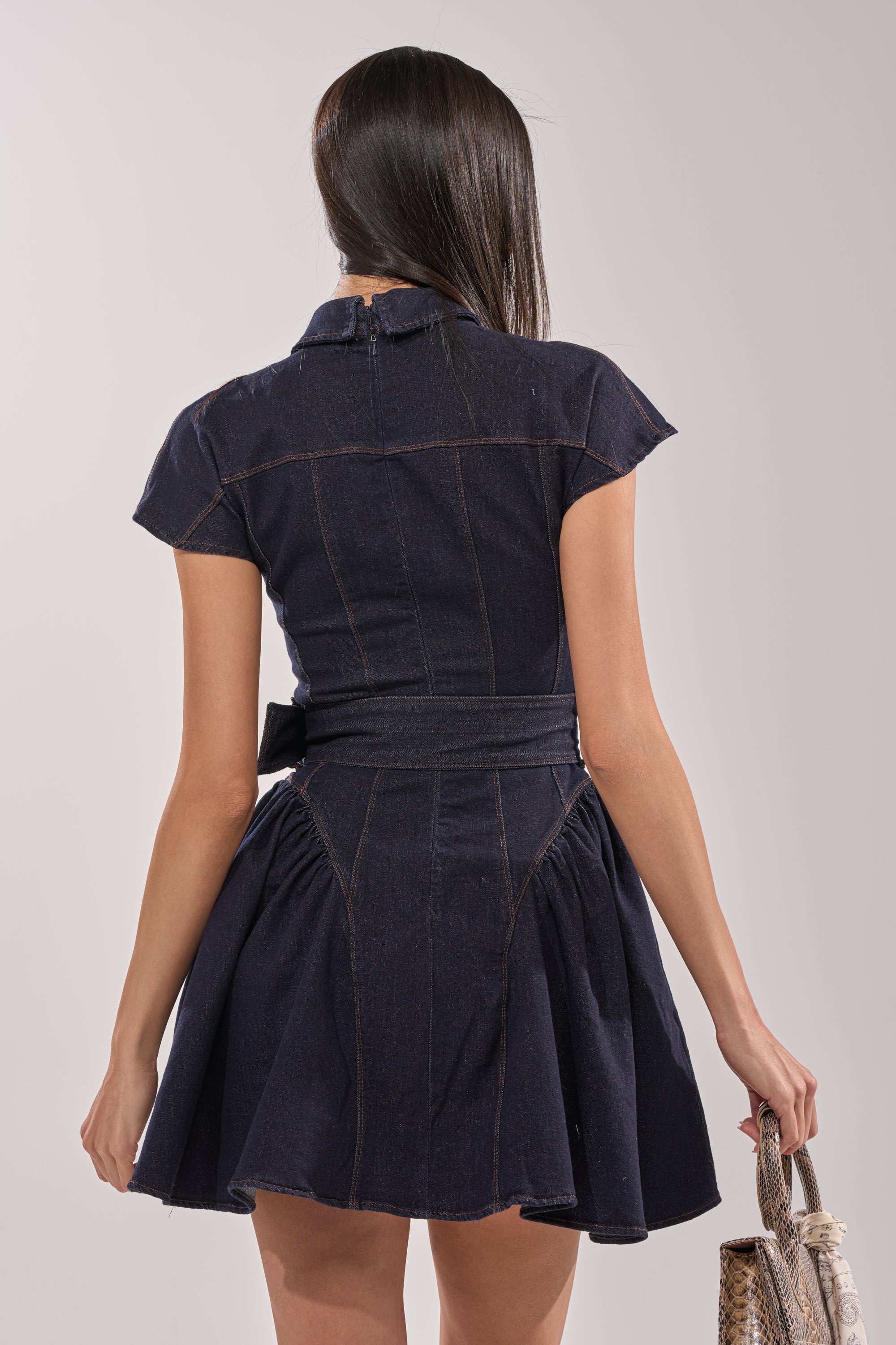 A woman with long, dark hair is seen from behind wearing the SADIE BUTTON UP DENIM MINI DRESS. The skater silhouette and belted waist are visible as she holds a textured handbag against a neutral background. 4