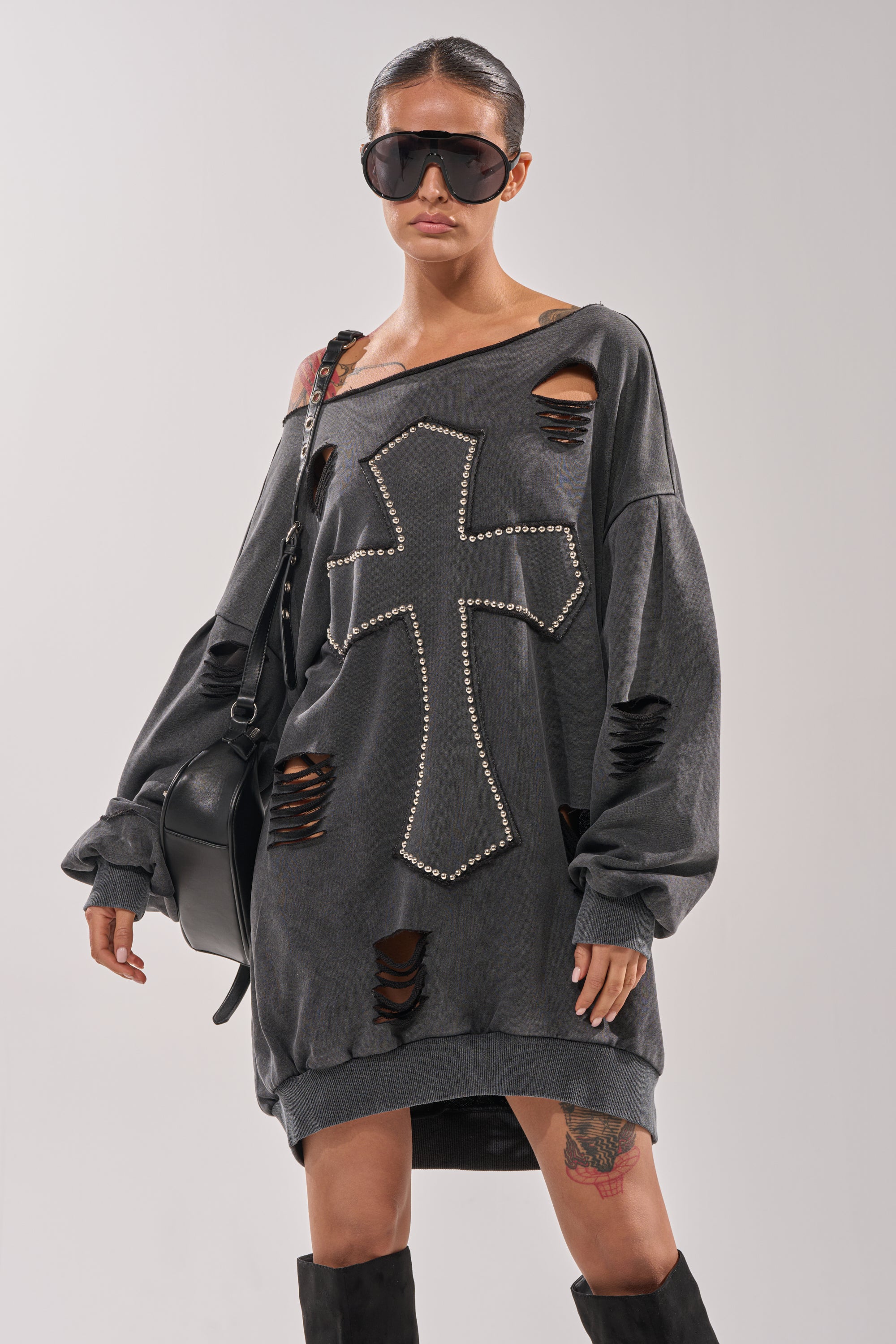 A woman in sunglasses wears the LEX STUDDED MINI SWEATSHIRT DRESS, an off-the-shoulder distressed black dress with a large cross, styled with a black shoulder bag, knee-high boots, slicked-back hair, and a visible leg tattoo. 0