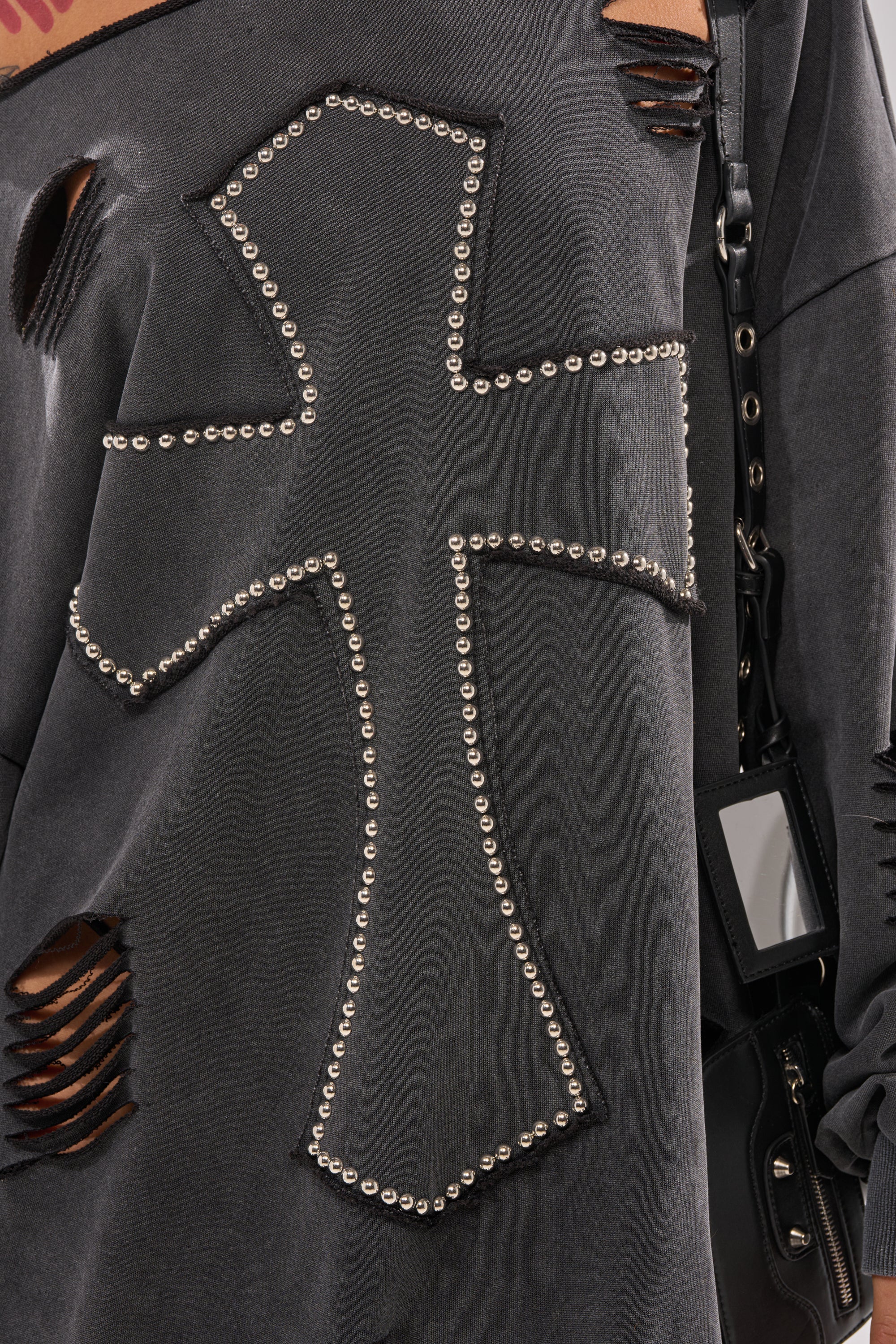A close-up of the LEX STUDDED MINI SWEATSHIRT DRESS, a distressed off-the-shoulder style with an oversized studded cross patch and intentional rips. A black bag with a rectangular tag hangs at the side. 9