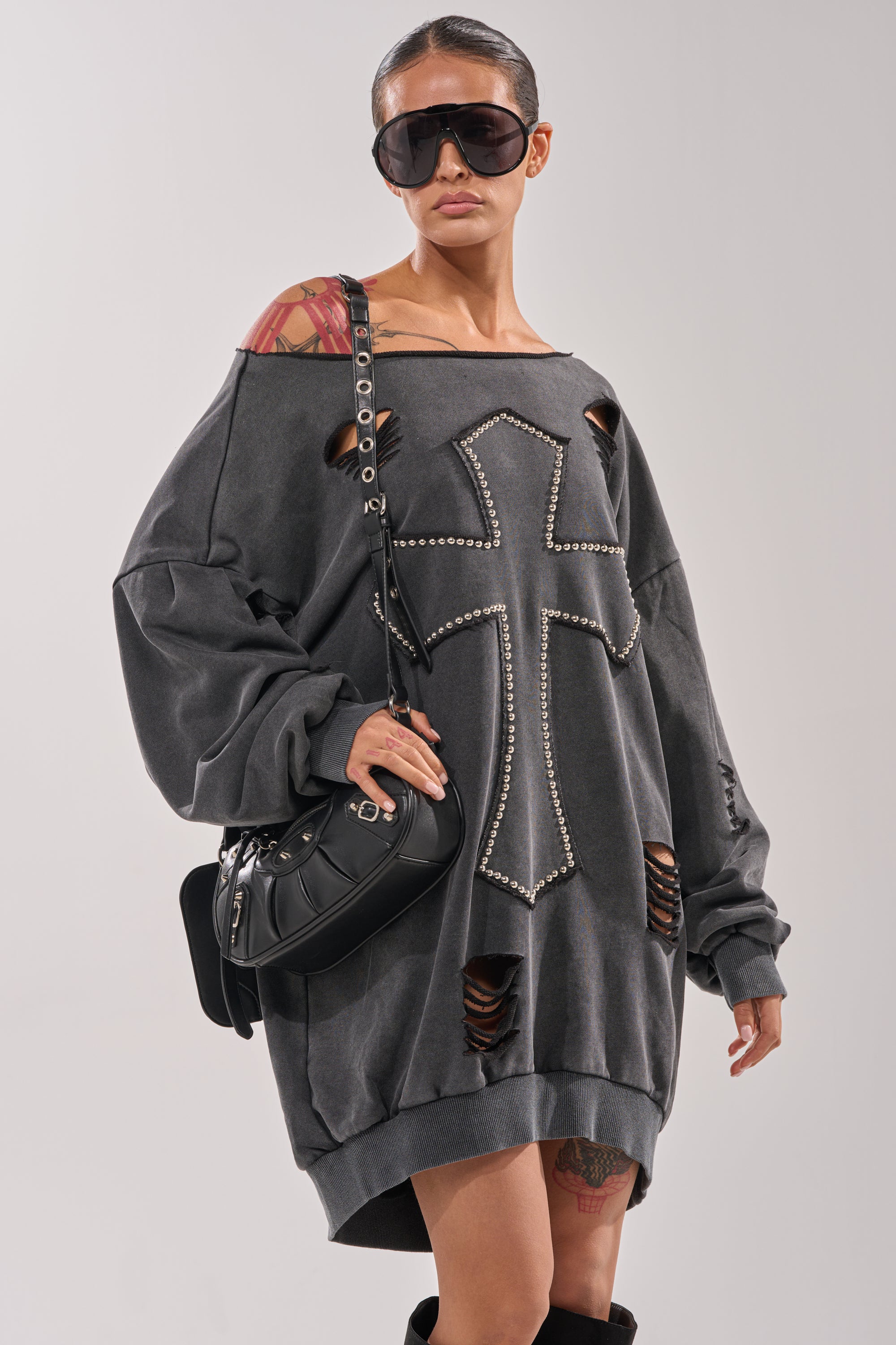 A woman in the LEX STUDDED MINI SWEATSHIRT DRESS, an off-the-shoulder cotton-blend piece with a bold oversized cross patch, pairs it with oversized sunglasses, a black handbag, and shows tattoos on her shoulder and thigh. 1