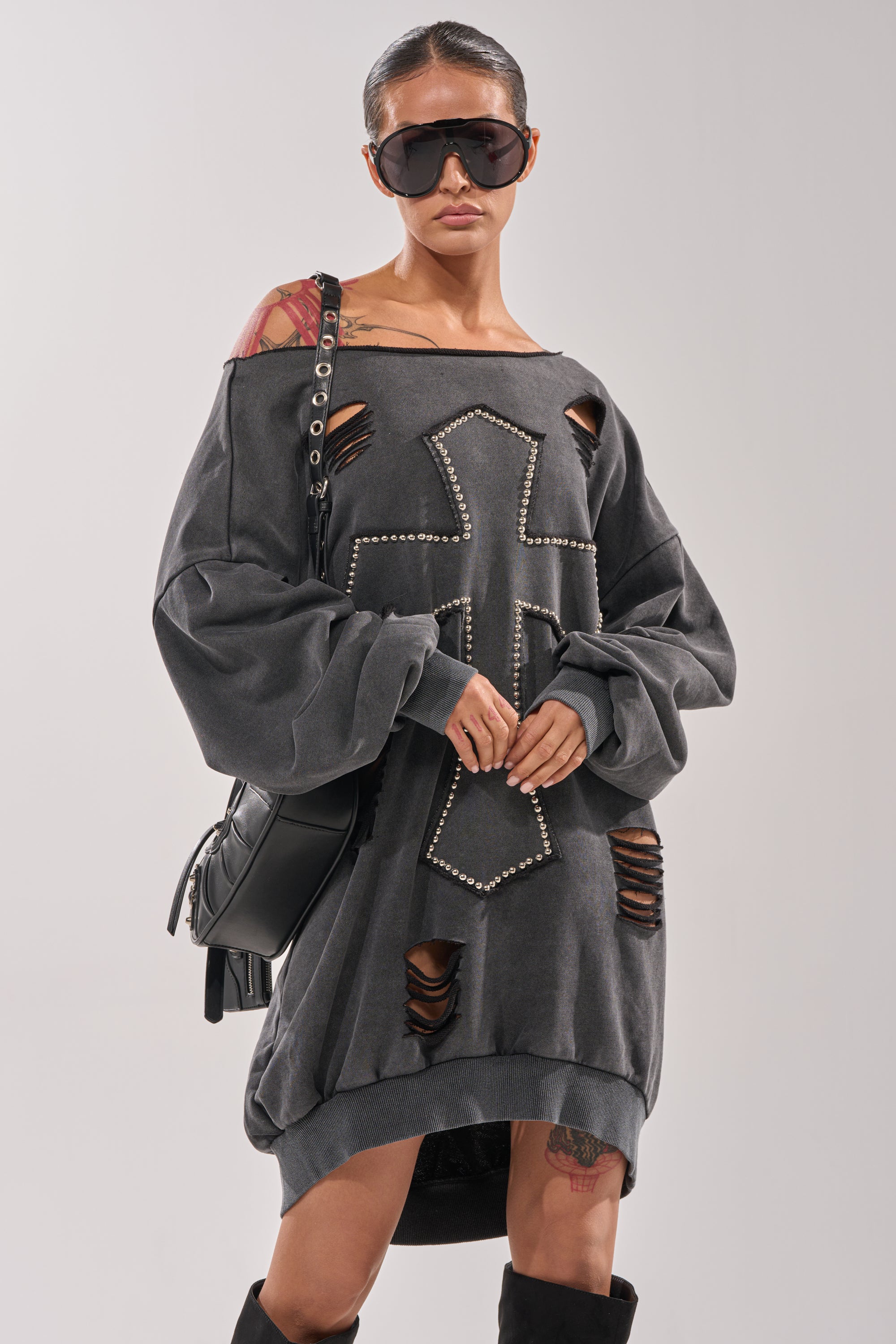 A woman stands confidently against a plain background wearing large black sunglasses, knee-high black boots, a black shoulder bag, and the LEX STUDDED MINI SWEATSHIRT DRESS—an oversized off-the-shoulder gray dress with a studded cross design. 2