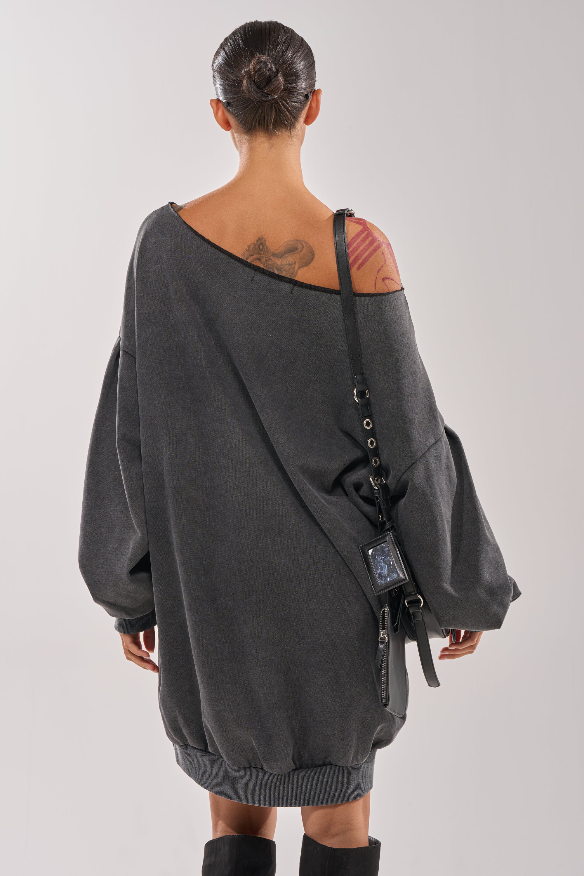 A person with dark hair in a bun, wearing the LEX Studded Mini Sweatshirt Dress off the shoulder, stands facing away. A shoulder tattoo is visible, and they carry a black crossbody phone case bag. 5