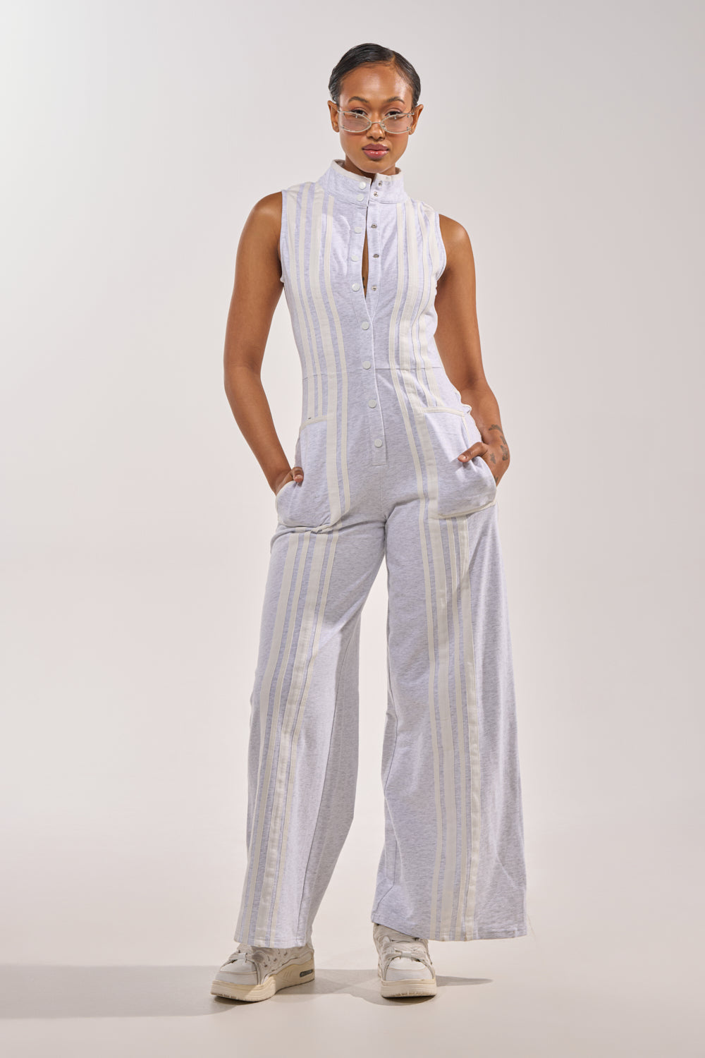 A person models the BIG SPORTS GIRL JUMPSUIT—sleeveless, cotton, with white vertical stripes and wide-leg pants—paired with white sneakers, standing against a plain light background with hands in pockets. 0