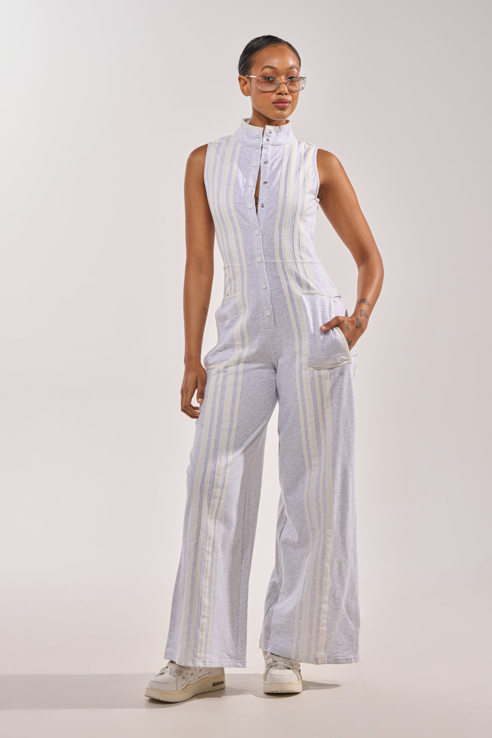A woman poses confidently in the BIG SPORTS GIRL JUMPSUIT—sleeveless cotton with vertical white and light gray stripes. She pairs it with white sneakers and glasses, her hair pulled back, standing against a plain background with one hand in her pocket. 2