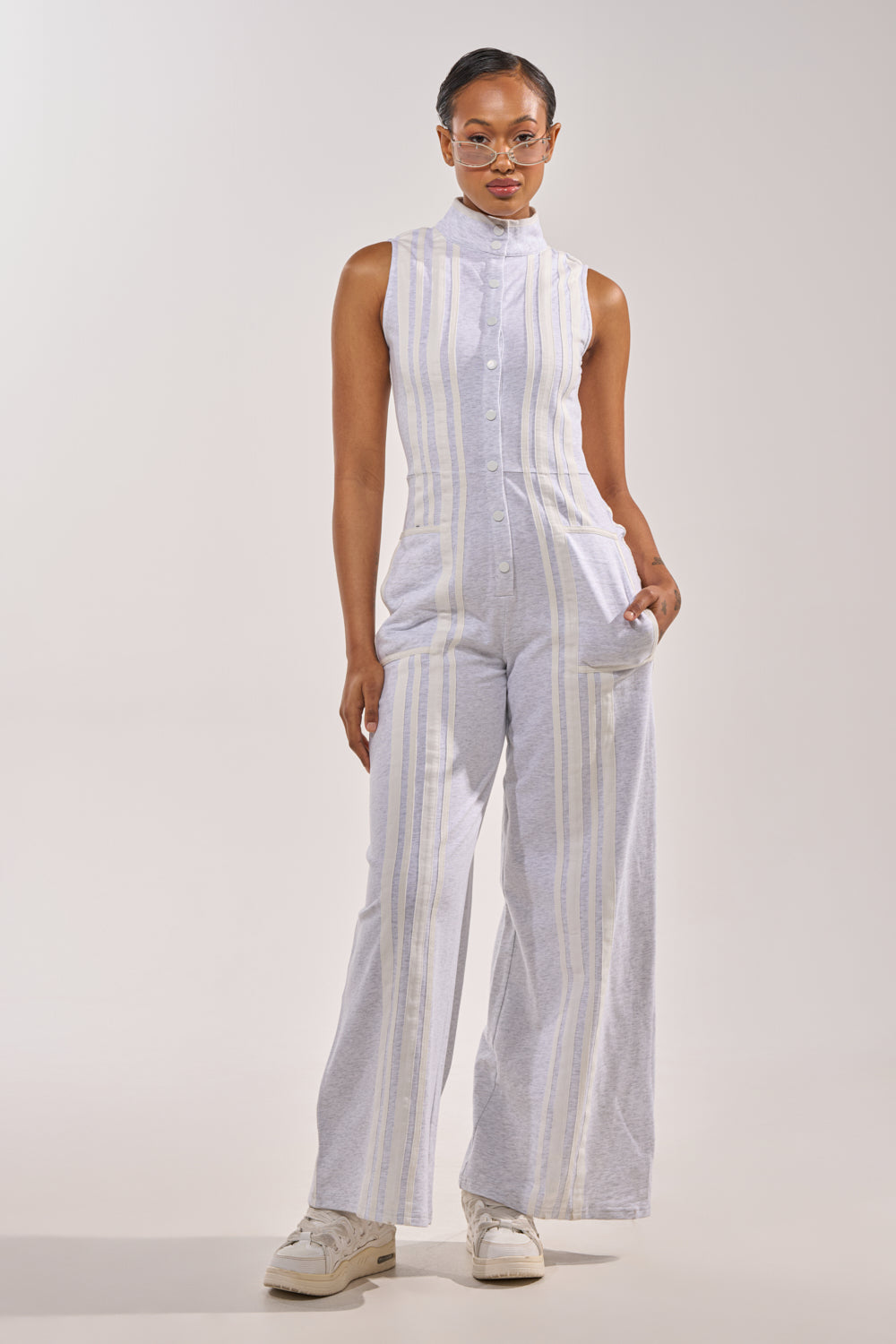 A person stands facing forward against a light gray background, wearing the BIG SPORTS GIRL JUMPSUIT—sleeveless with vertical pastel stripes and wide-leg pants—paired with white sneakers and clear glasses, one hand in a pocket. 3