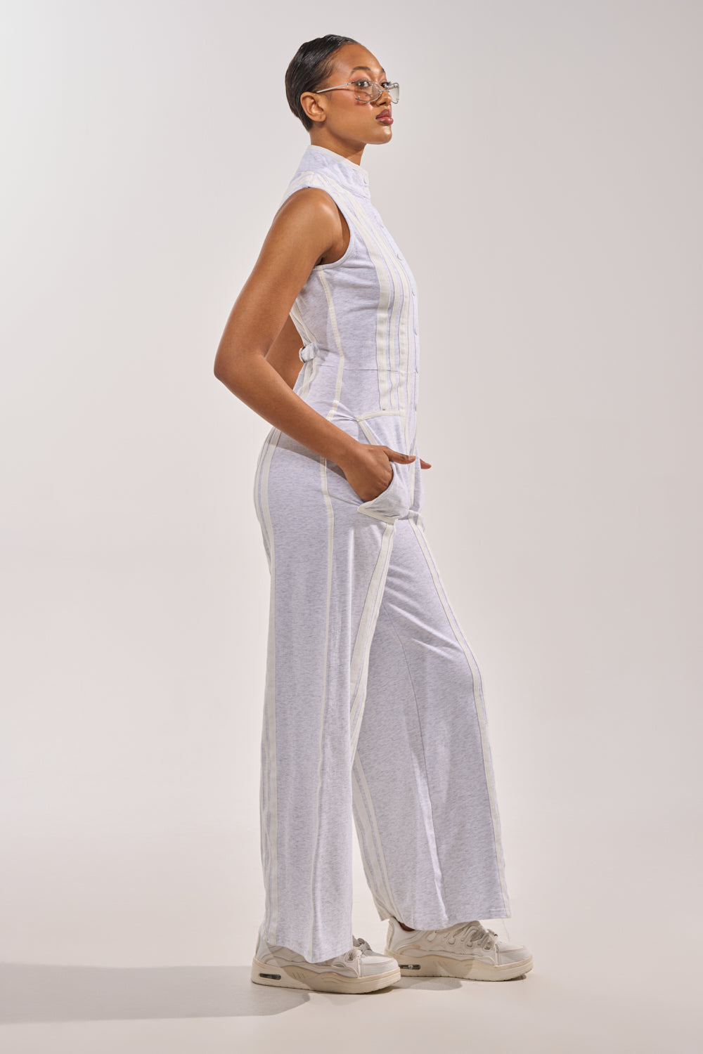 A person in profile stands against a light background, wearing the BIG SPORTS GIRL JUMPSUIT—sleeveless with a high collar, wide legs, and pockets—paired with white sneakers and clear glasses. 4