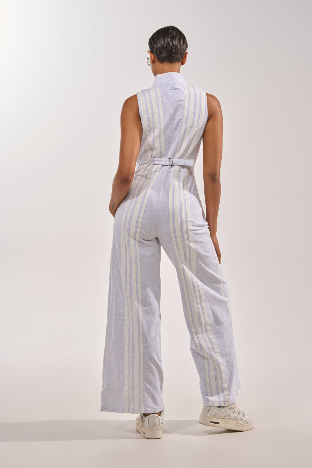 A person with short hair wears the BIG SPORTS GIRL JUMPSUIT, a sleeveless cotton one-piece with vertical stripes and a belted waist, paired with white sneakers against a plain background. 5