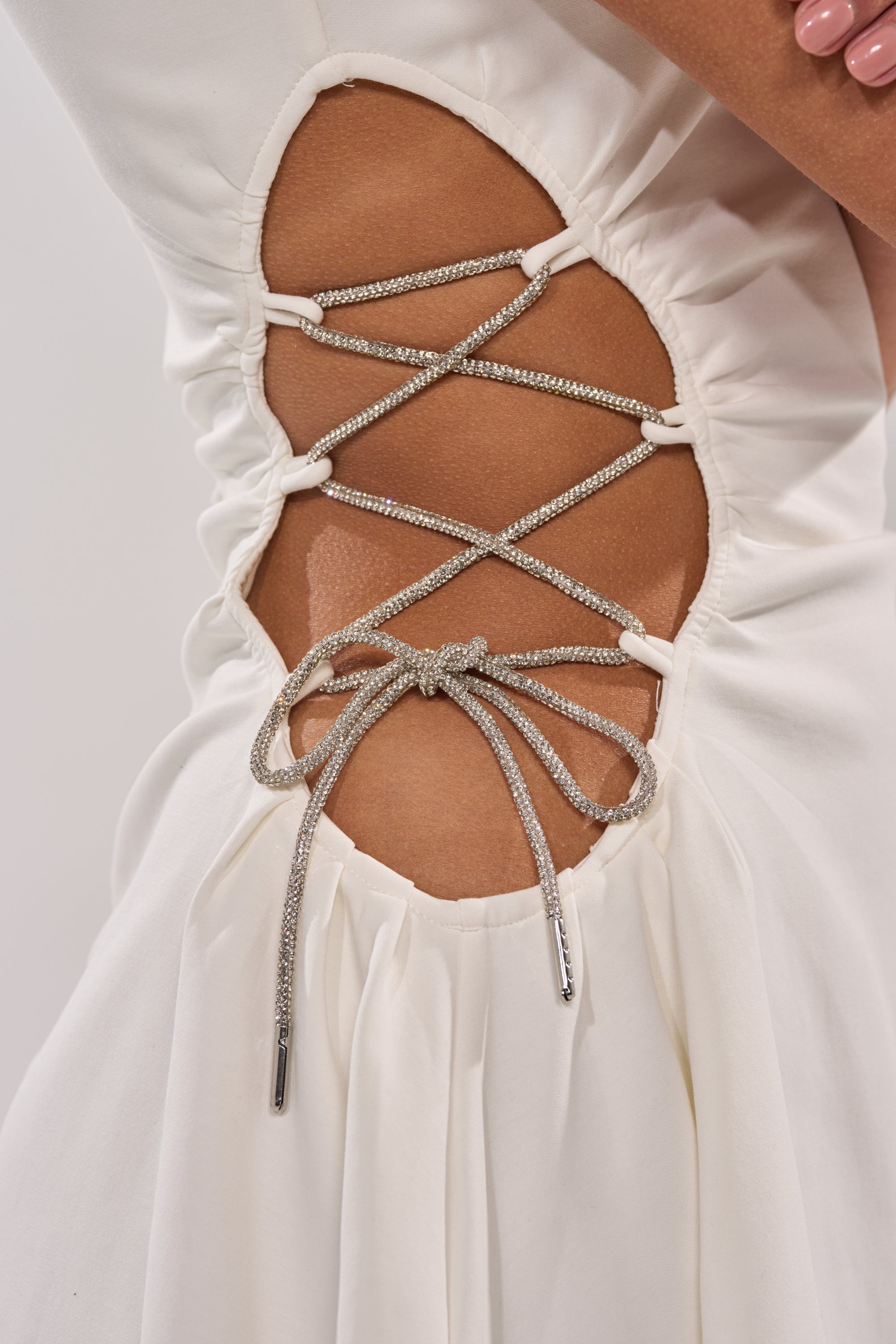 Close-up of the STAY CLASSY CUT OUT MINI DRESS—a white mini dress with a side cut-out, silver lace-up string detail revealing skin beneath the crisscrossed laces; a person’s arm with light pink nails appears in frame. 9