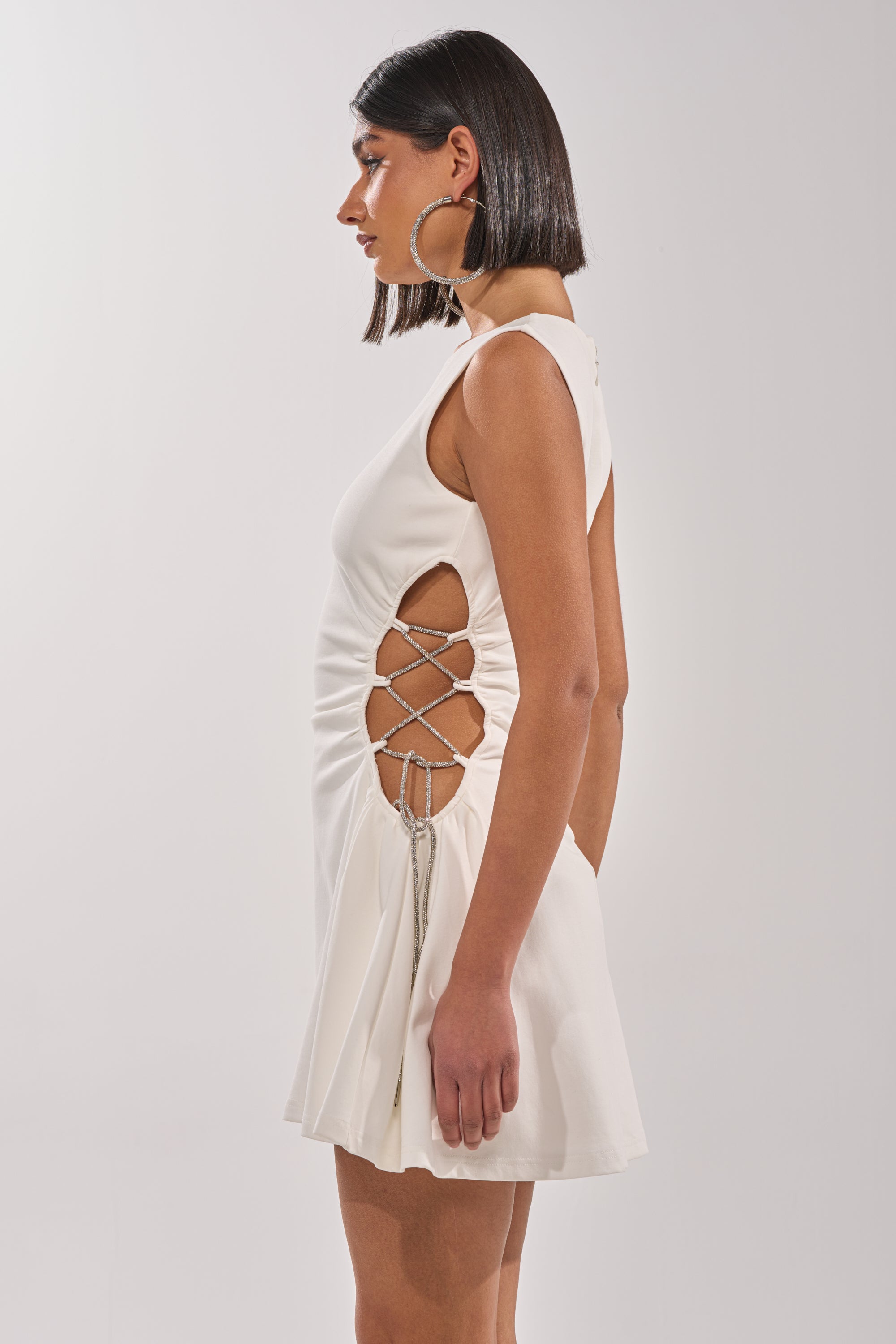 A woman stands in profile against a plain light background, wearing the STAY CLASSY CUT OUT MINI DRESS with side lace-up cutouts. She completes her summer party look with large hoop earrings and straight dark hair. 3