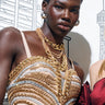 A person wearing a glamorous dress, accessorized with the OMG Slay Layered Chain Necklace in Gold and sparkling jewelry, stands against a backdrop featuring illustrated cityscape elements while holding a metallic clutch.