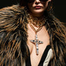 A fashion model showcases the LOVER LAYERED NECKLACE, featuring multiple gold chains with a bold Cuban chain and a striking jeweled cross pendant, while wearing large sunglasses and a fur coat. The dramatic deep neckline of her outfit, adorned with ornate buttons, stands out against the dark background.