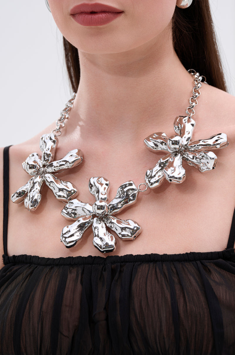 A woman wears a sheer black top and the BELLI NECKLACE—a chunky silver statement piece with three large, textured flower pendants. The image is cropped to show her from the shoulders up.