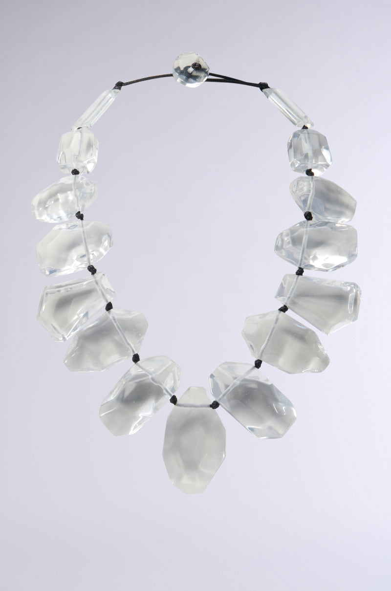The CLEARLY CHIC NECKLACE features large translucent stones linked by a thin black cord, with small black crystal beads between each stone, exuding understated elegance against a plain grey backdrop.
