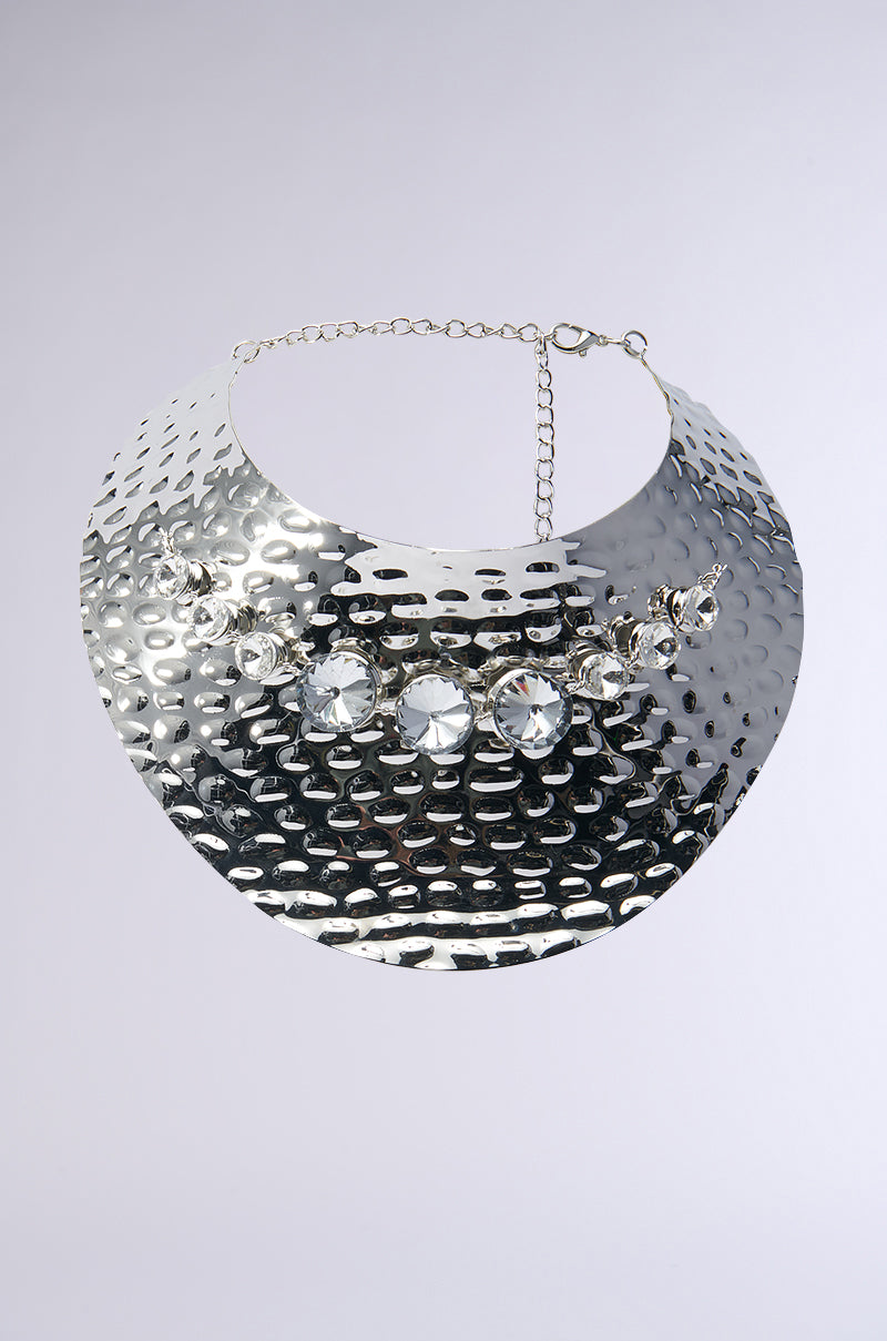 The CANT BREAK ME TEXTURED METAL NECKLACE is a shiny, hammered silver piece with an adjustable clasp and features large, reflective gemstones arranged in a row along the middle.