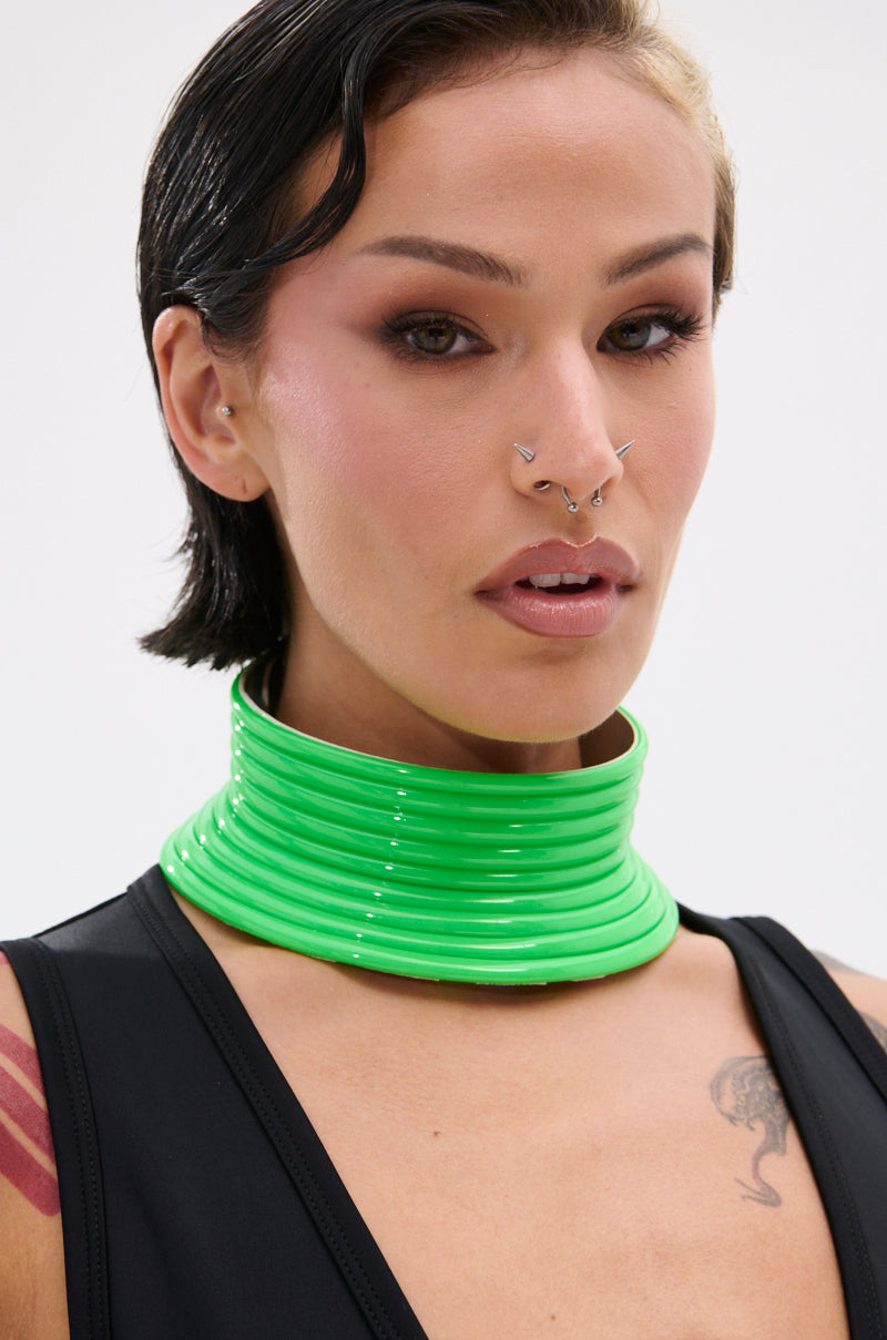 A person with short dark hair, facial piercings, and tattoos wears a black top and the TOO BRAT NECKLACE—a chunky neon green statement piece—adding futuristic flair as they look directly at the camera.