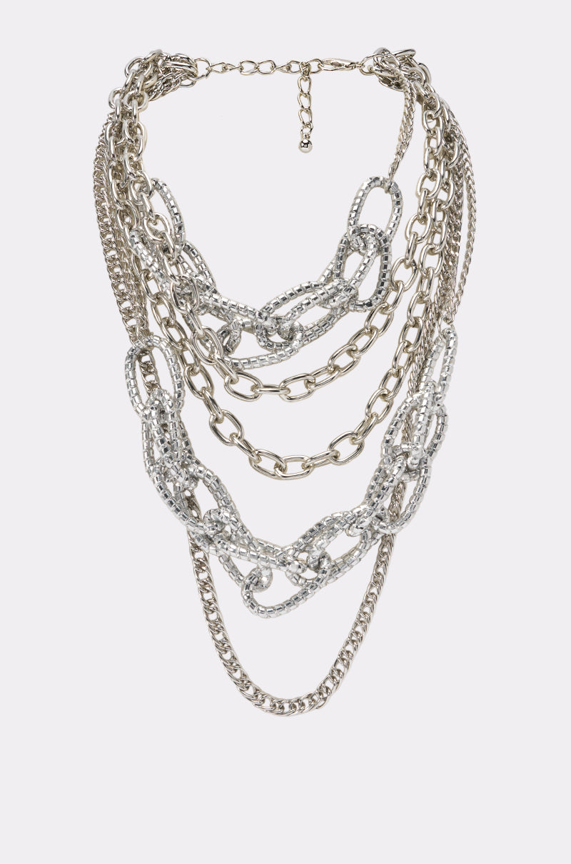 Introducing the LAYERS ON LAYERS NECKLACE: a striking multi-layered statement piece crafted with mixed silver chains in a variety of links and textures, from bold thick strands to delicate thin ones, some adorned with sparkling rhinestone embellishments. Presented on a plain white background, this eye-catching silver-tone necklace effortlessly combines modern edge with timeless elegance.