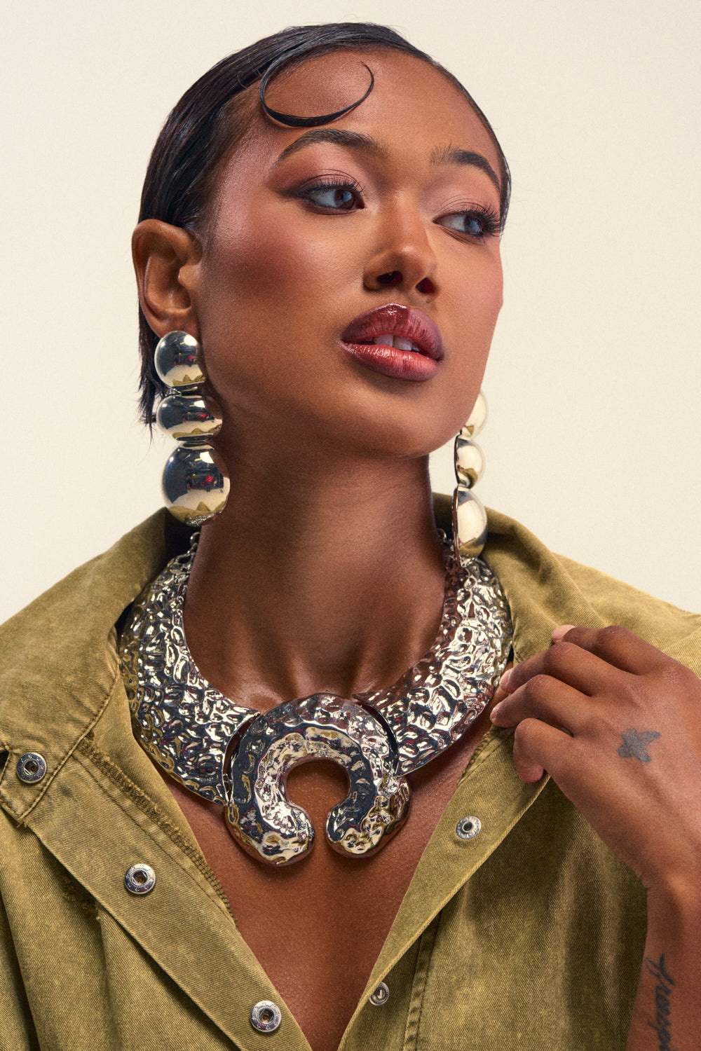 Wearing the WYM NECKLACE—a bold, hammered silver statement piece—a woman pairs her contemporary look with large metallic earrings and a khaki shirt. With slicked-back hair and a small star tattoo on her hand, she exudes confidence as she gazes to the side.