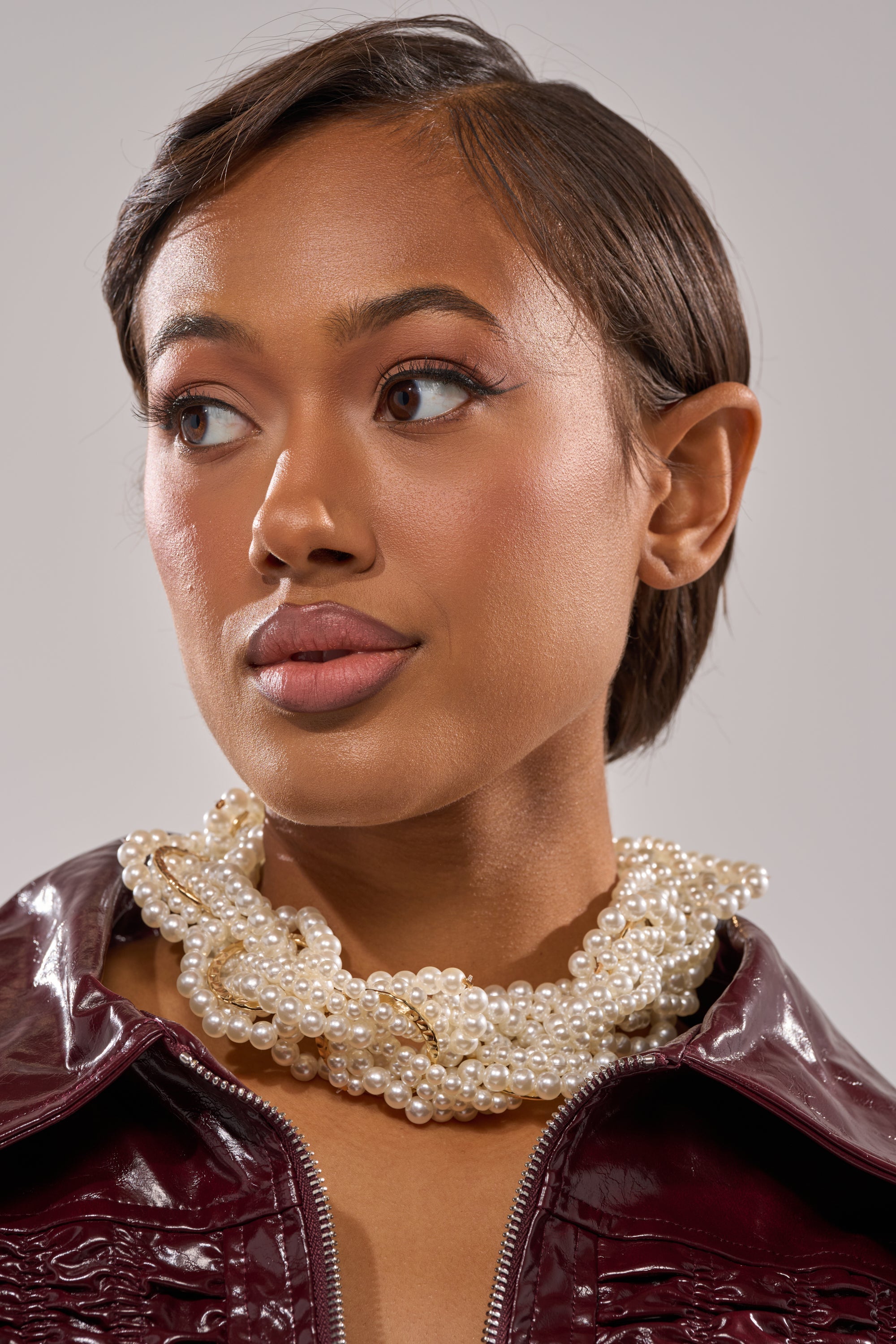 A woman with short brown hair wears a burgundy leather jacket and the SHARON NECKLACE, featuring gold-tone accents, while looking slightly to her left against a neutral background. 1