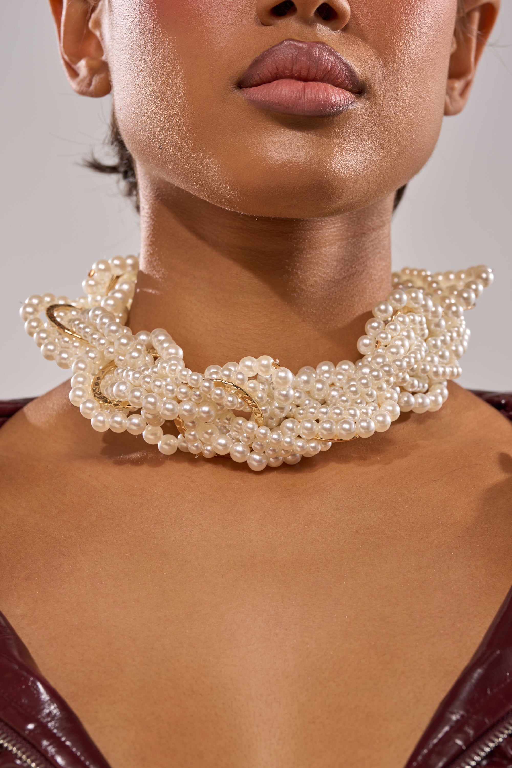 A woman with medium brown skin and a burgundy top showcases the SHARON NECKLACE, a twisted multi-strand gold-tone piece accented with faux pearls, drawing attention to her elegant neck and jewelry. 2