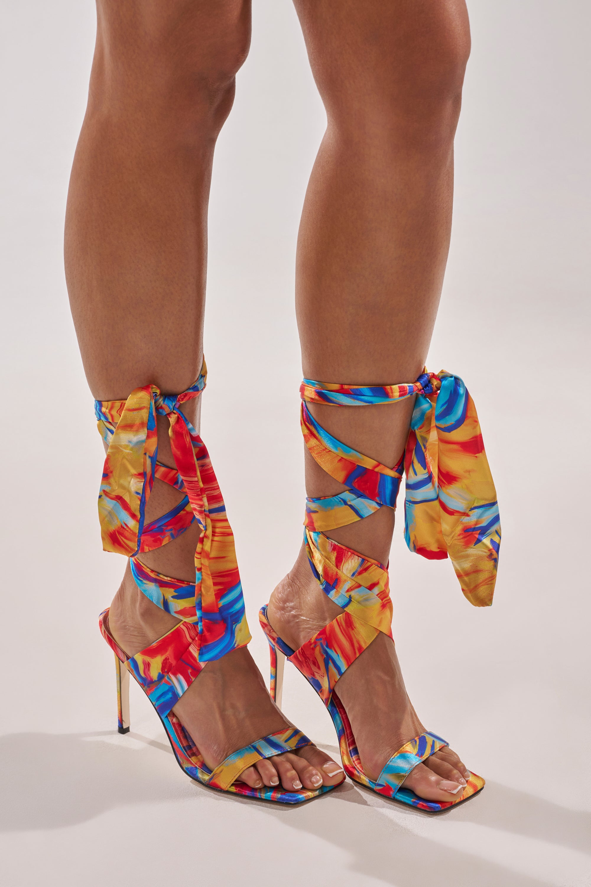 Someone wears the AZALEA WANG JOHA ORANGE MULTI PRINTED SATIN WRAP UP STILETTO, featuring vibrant blue, red, and yellow patterns, open square toes, and colorful satin straps that wrap around the ankles and tie in bows. 0
