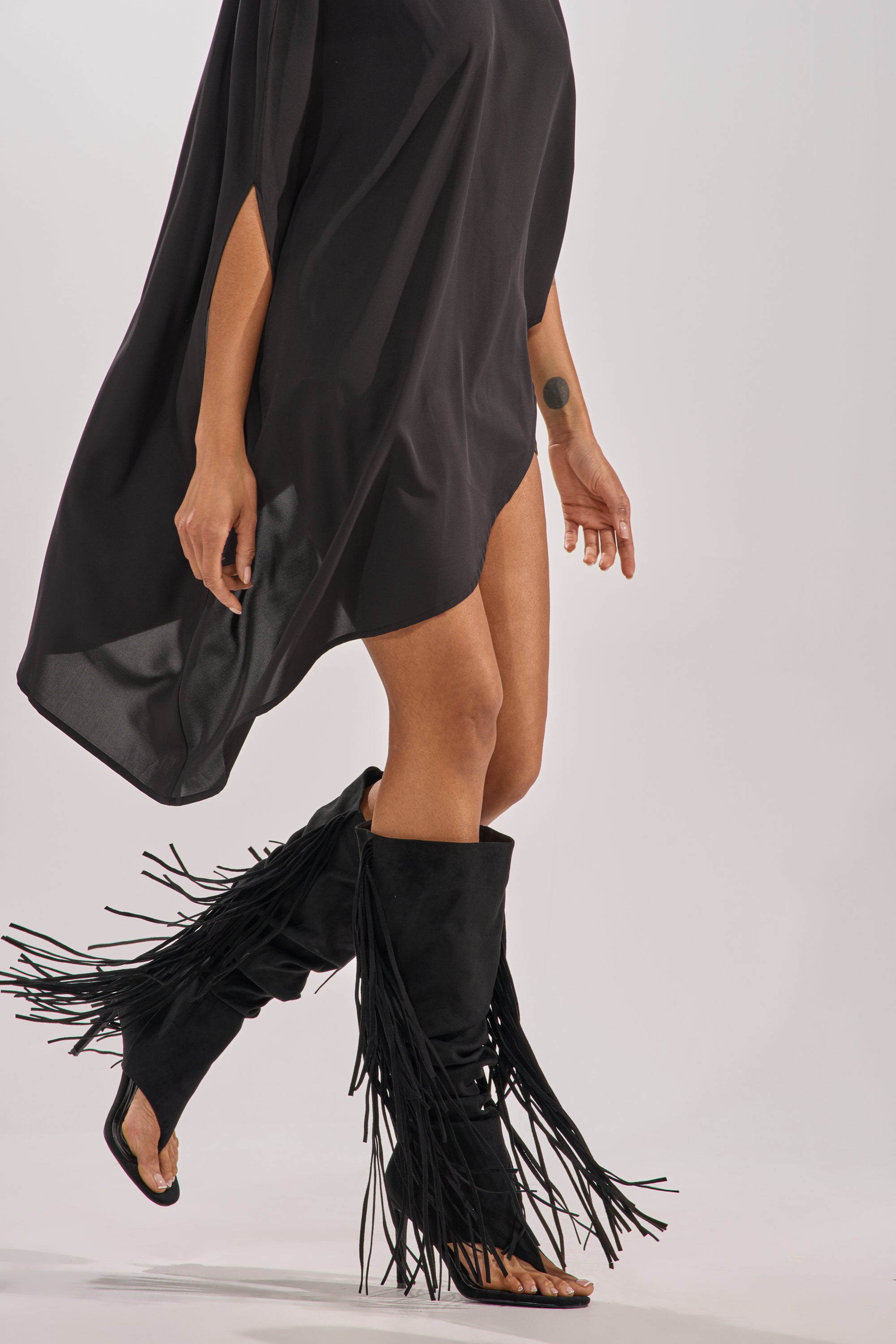 A person in a flowing black dress walks forward, showing their legs and wearing the AZALEA WANG JOHANNA BLACK FRINGE GLADIATOR SANDAL with stiletto heels. The image is cropped to display only the lower half of their body. 2