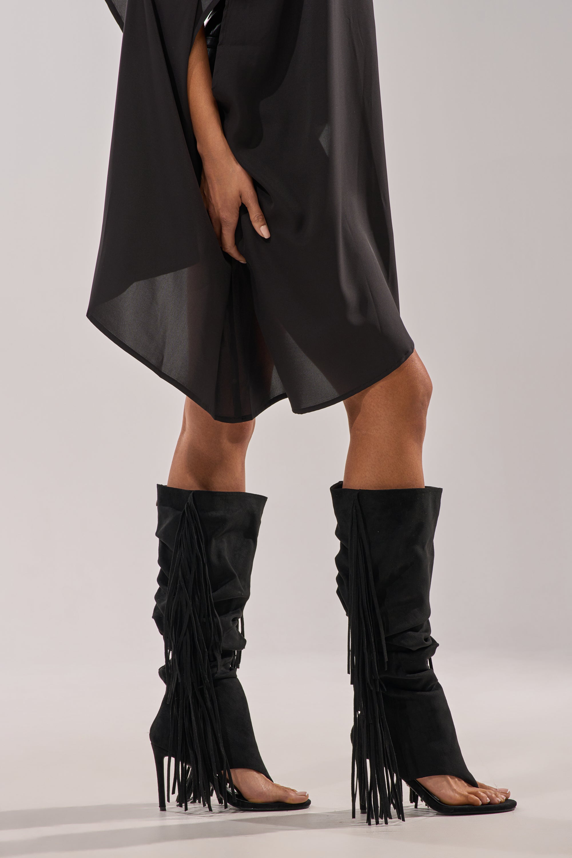 A person stands on a light surface wearing a sheer black dress and AZALEA WANG JOHANNA BLACK FRINGE GLADIATOR SANDALS. The image is cropped from the shoulders down. 3