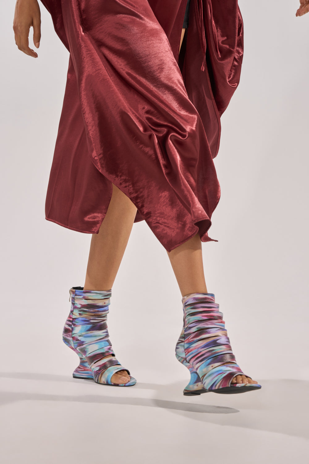 A person in a shiny maroon skirt and AZALEA WANG JOHANNES MULTI RAINBOW WEDGE OPEN TOE BOOTIES with wavy, sculpted soles walks against a neutral background. 1