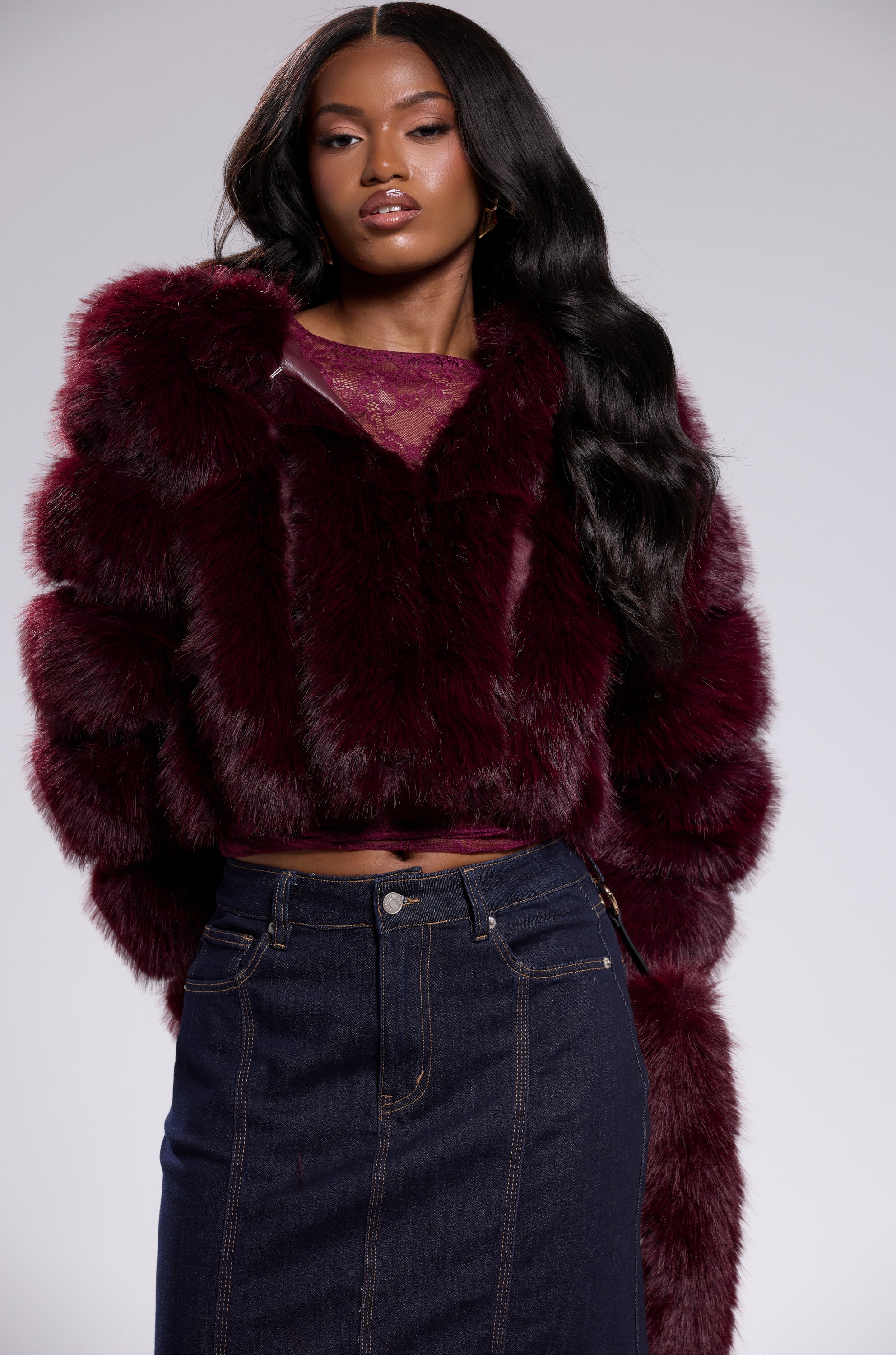 A woman with long, wavy hair confidently poses in the JOLENE FAUX FUR COAT IN BURGUNDY over a lace top and dark denim skirt, standing against a plain light background.