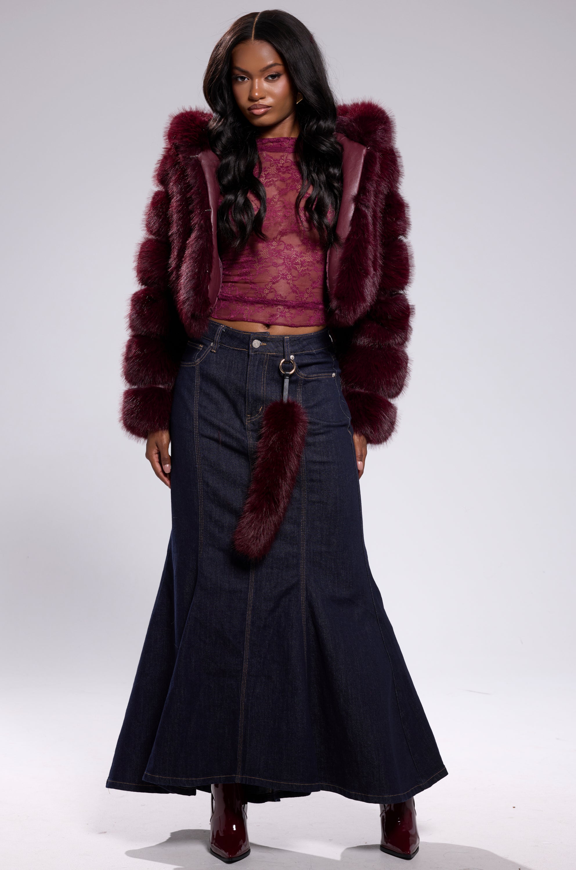 A woman wears the JOLENE FAUX FUR COAT IN BURGUNDY over a sheer lace top, a long denim skirt with a fur-tail belt, and burgundy boots, standing against a light gray background.