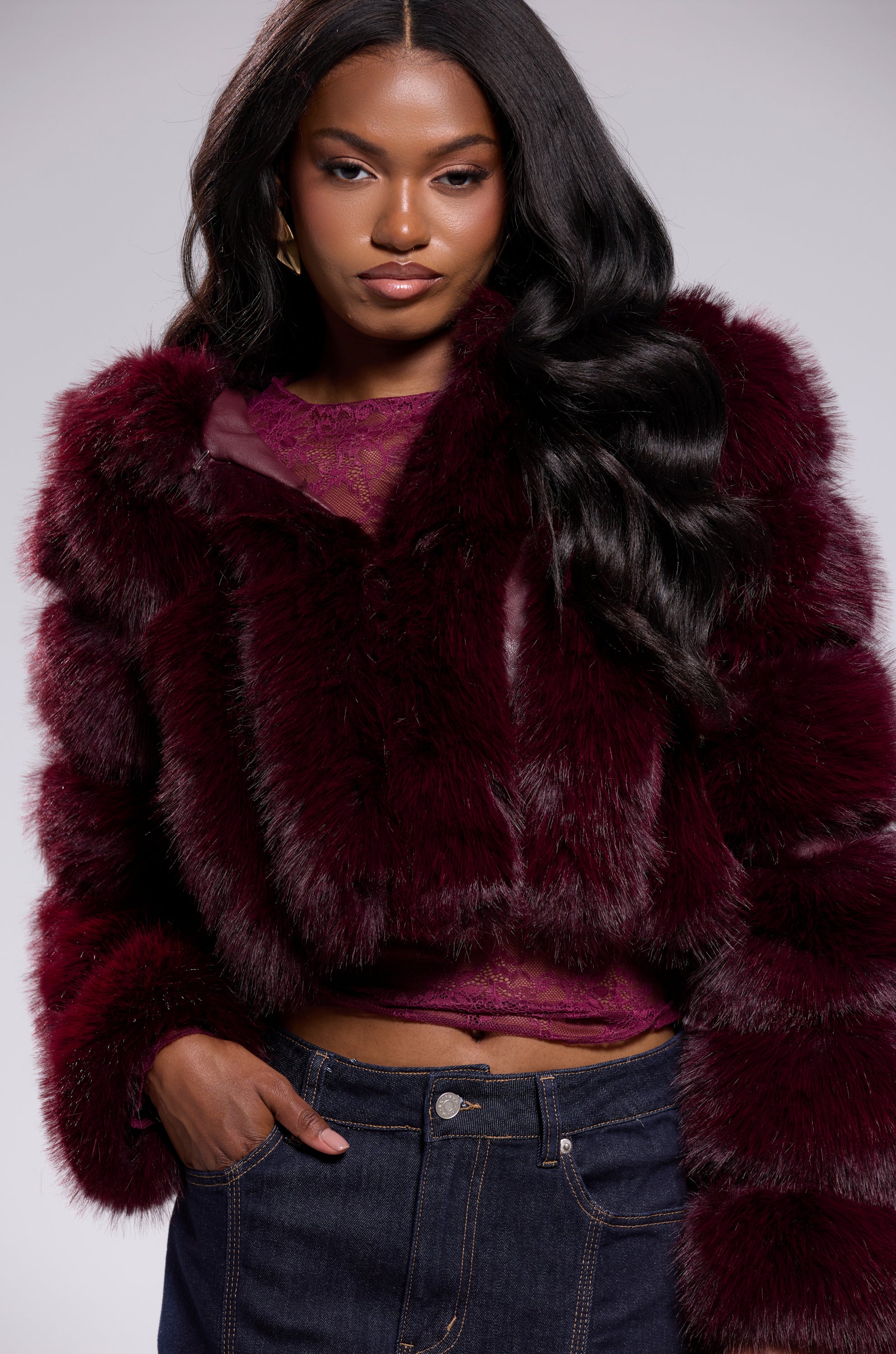 A woman with long, wavy black hair models the JOLENE FAUX FUR COAT IN BURGUNDY over a lace top and dark jeans, posing confidently against a plain light gray background.