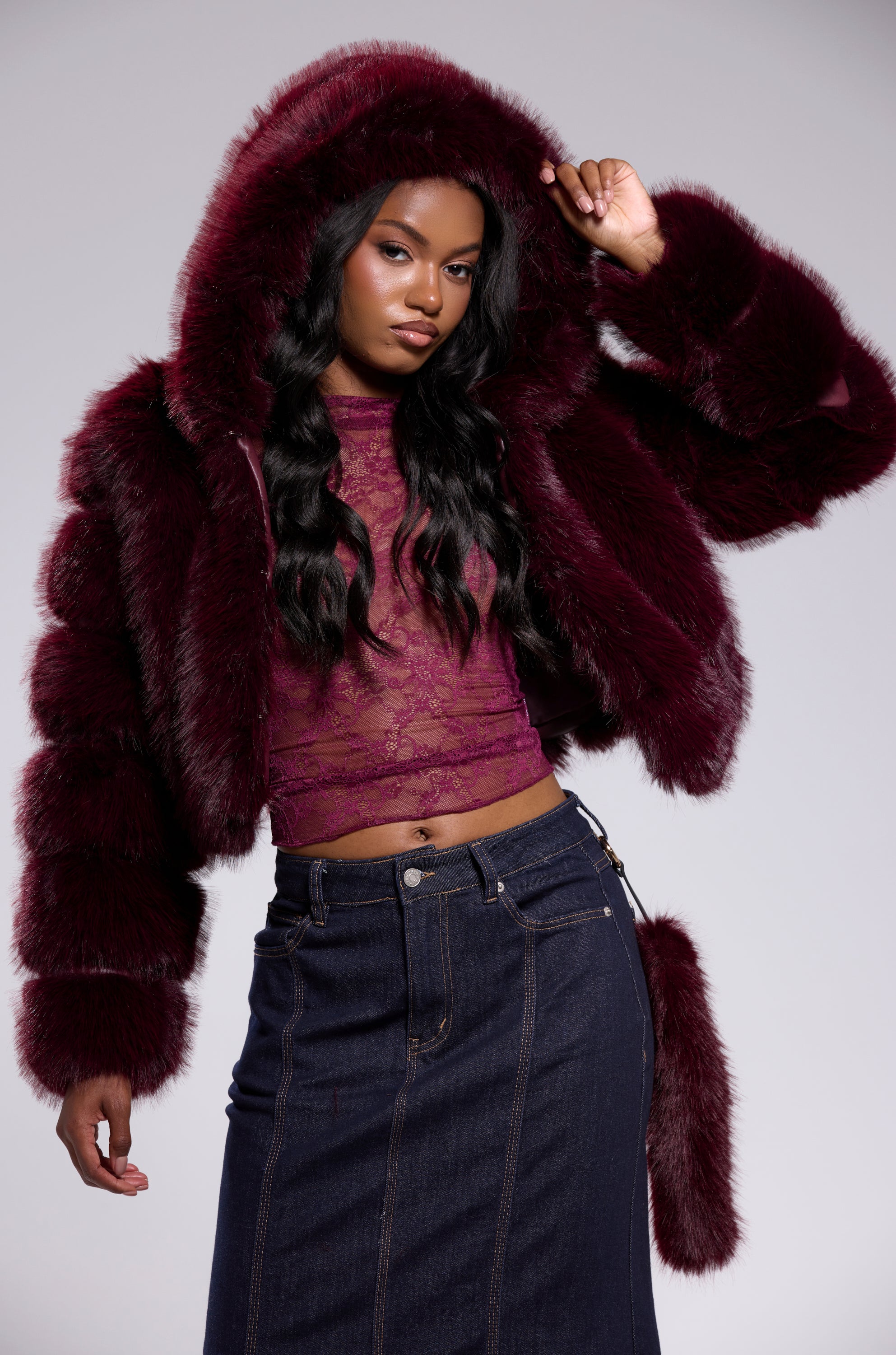 A woman models the JOLENE FAUX FUR COAT IN BURGUNDY with a cropped cut, sheer lace top, and dark denim skirt. She lifts the hood, looking confidently at the camera against a plain backdrop.