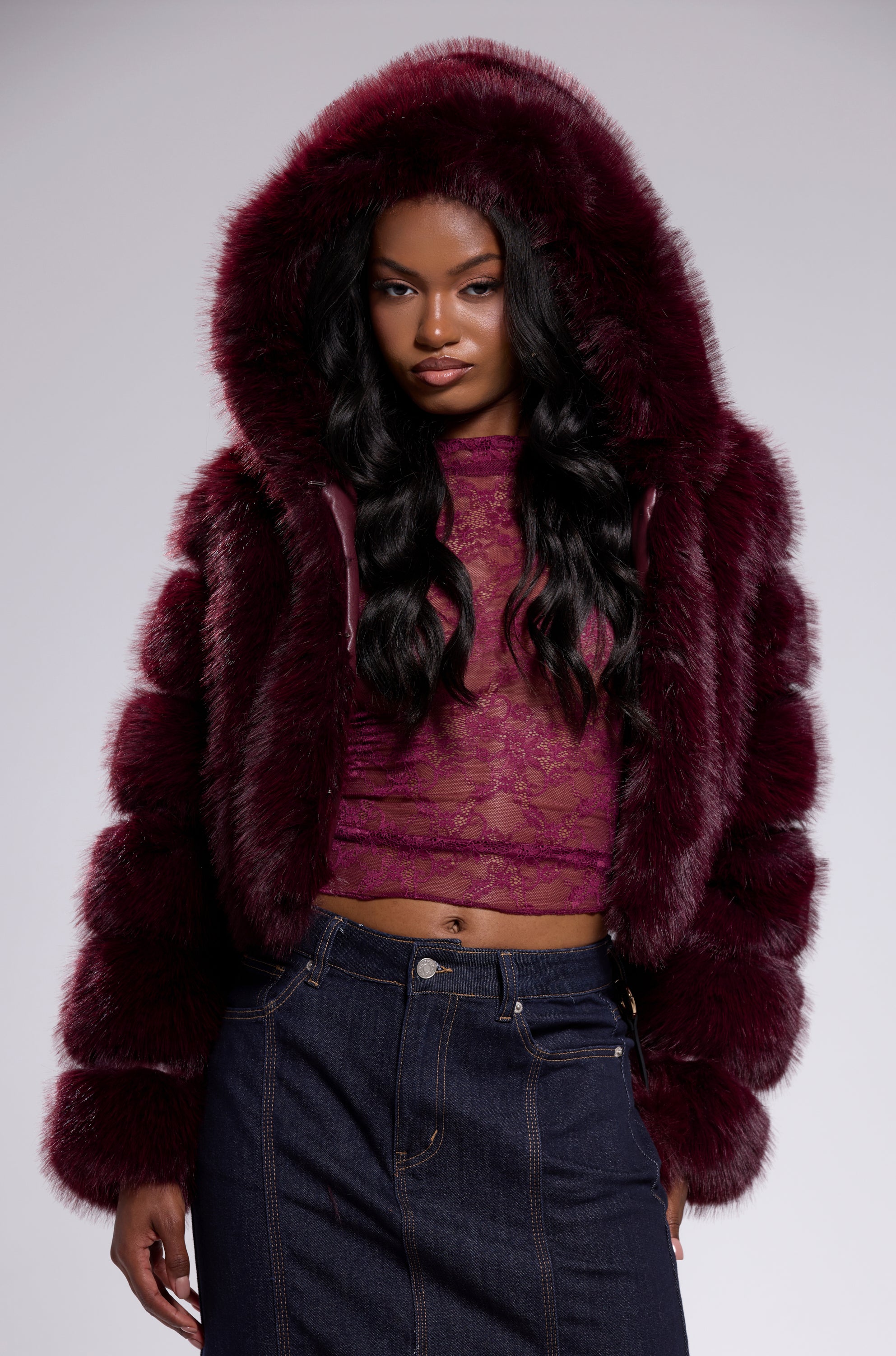 A woman in the JOLENE FAUX FUR COAT IN BURGUNDY with a hood stands facing forward over a sheer lace top and dark denim skirt against a neutral background.