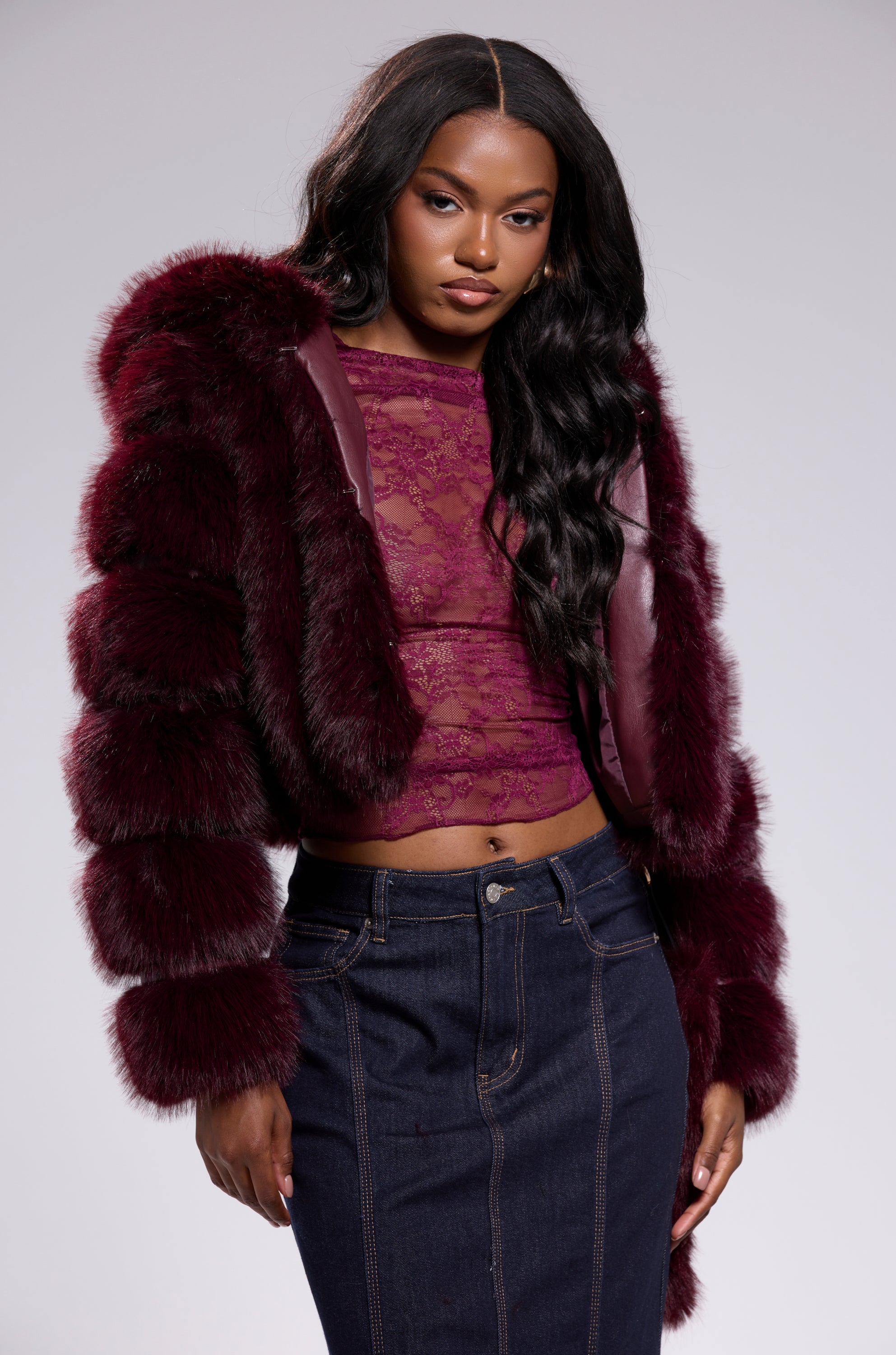 A woman stands confidently against a light gray background, wearing the JOLENE FAUX FUR COAT IN BURGUNDY over a matching lace top and dark denim skirt.
