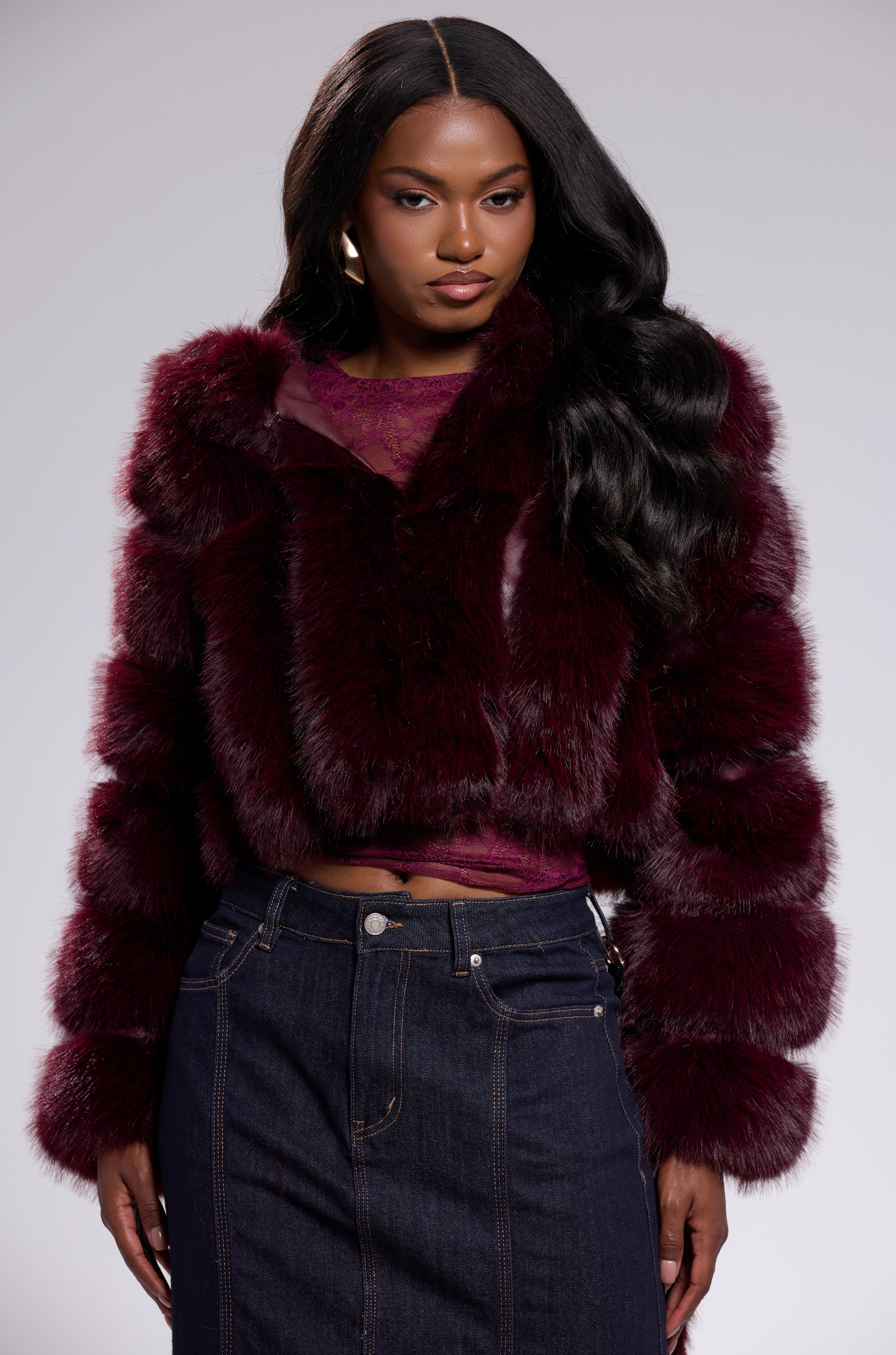 A woman with long wavy hair wears the JOLENE FAUX FUR COAT IN BURGUNDY over a sheer burgundy top and blue high-waisted jeans, standing against a plain light background.