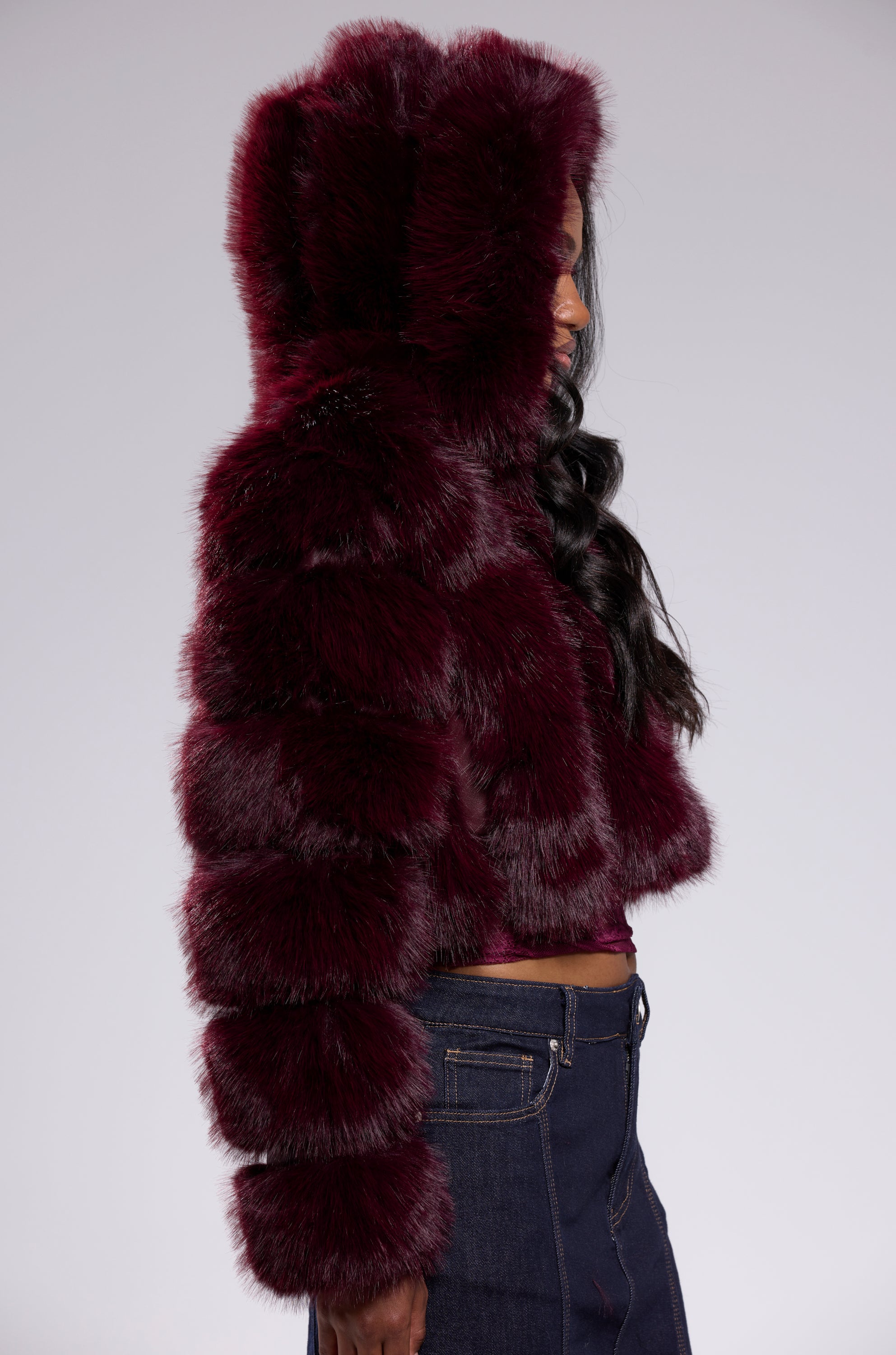 A person wears the JOLENE FAUX FUR COAT IN BURGUNDY styled as a cropped jacket with dark blue high-waisted jeans, shown from the side against a plain light background.
