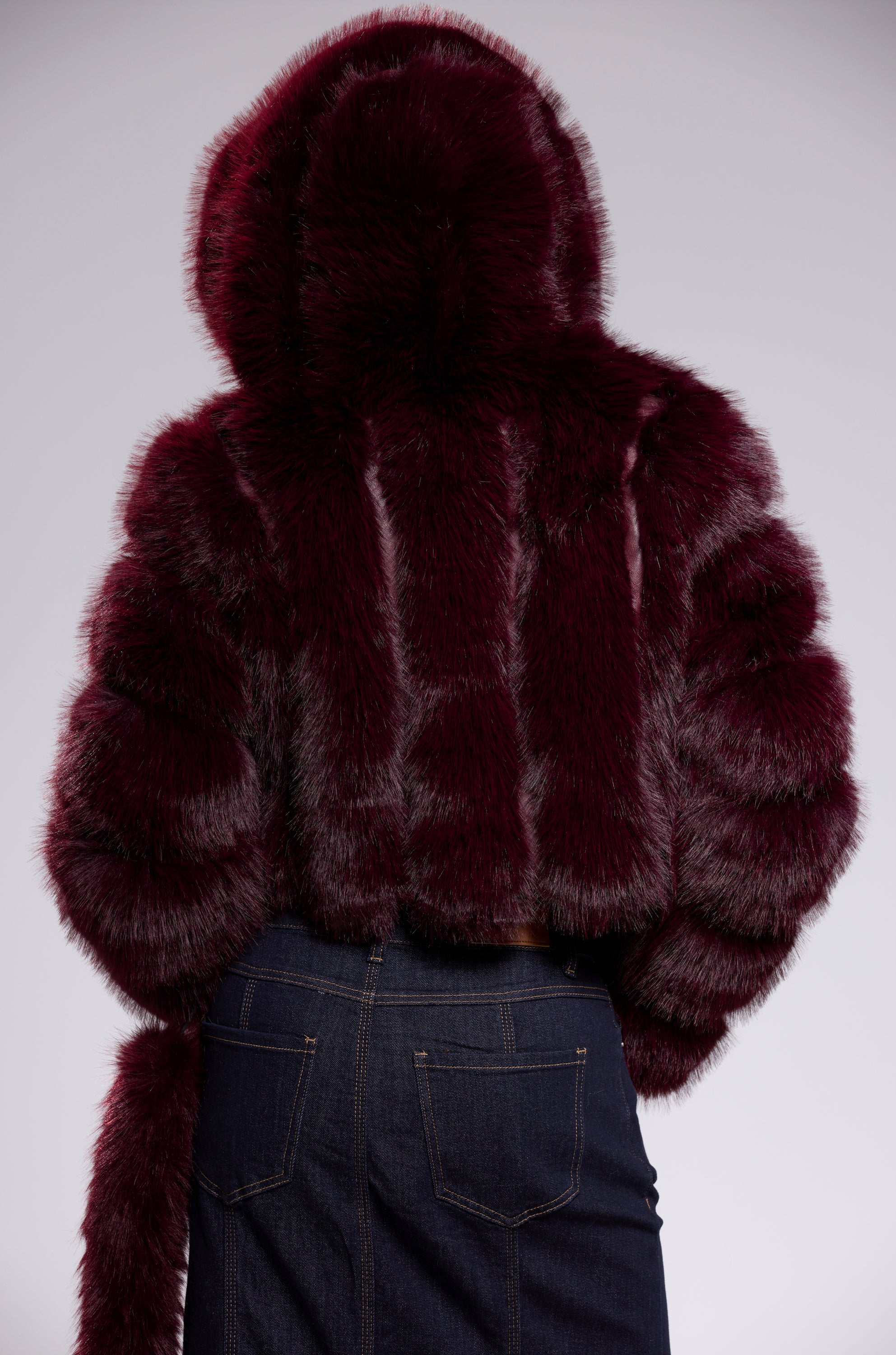 A person is seen from the back wearing the JOLENE FAUX FUR COAT IN BURGUNDY, featuring a hooded, striped-texture and cropped silhouette, paired with dark denim jeans against a plain light background.