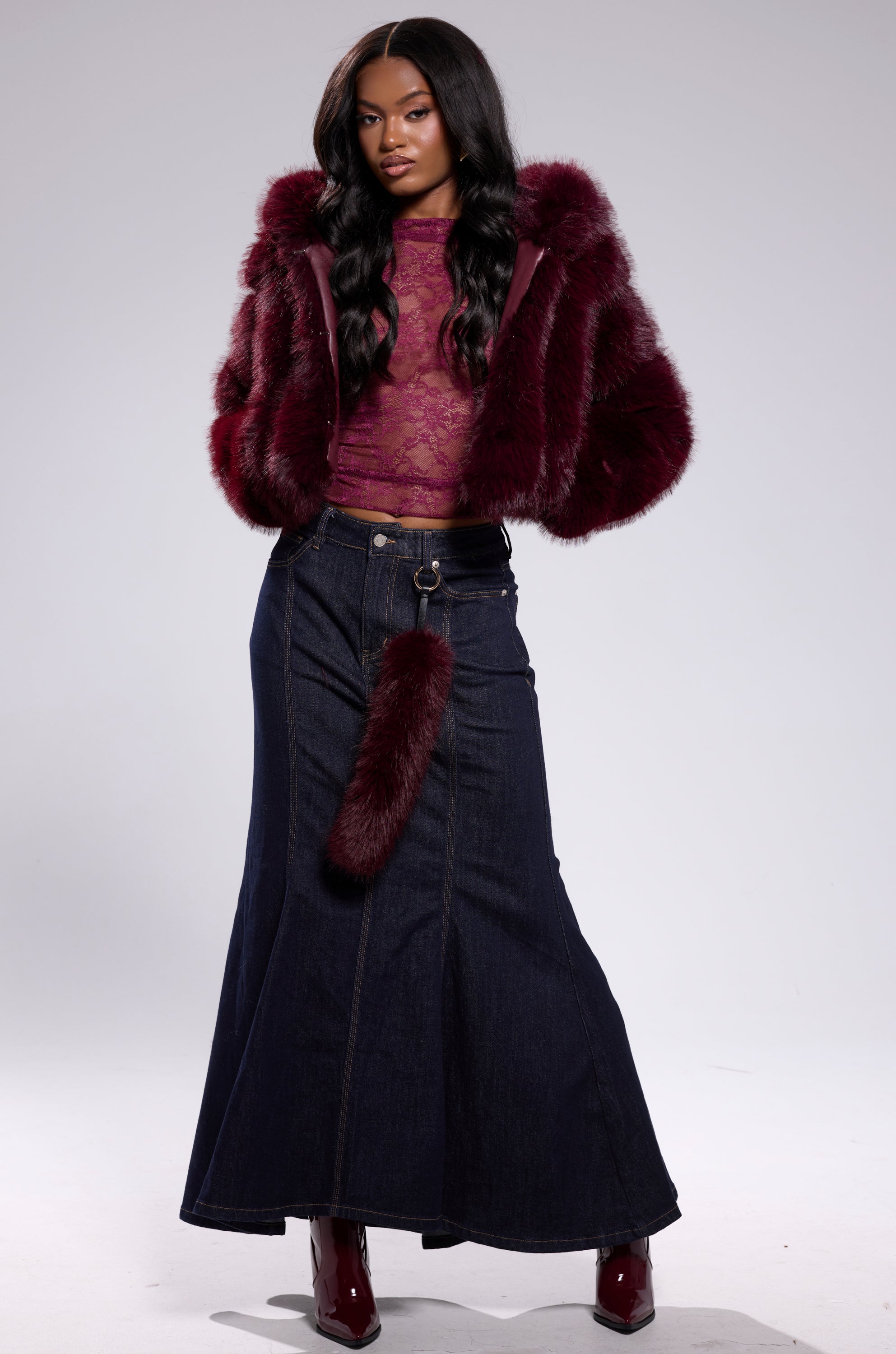 A woman poses against a plain background in the JOLENE FAUX FUR COAT IN BURGUNDY, styled over a sheer lace top, a long dark denim skirt, matching burgundy heeled boots, and a furry accessory hanging from her waistband.